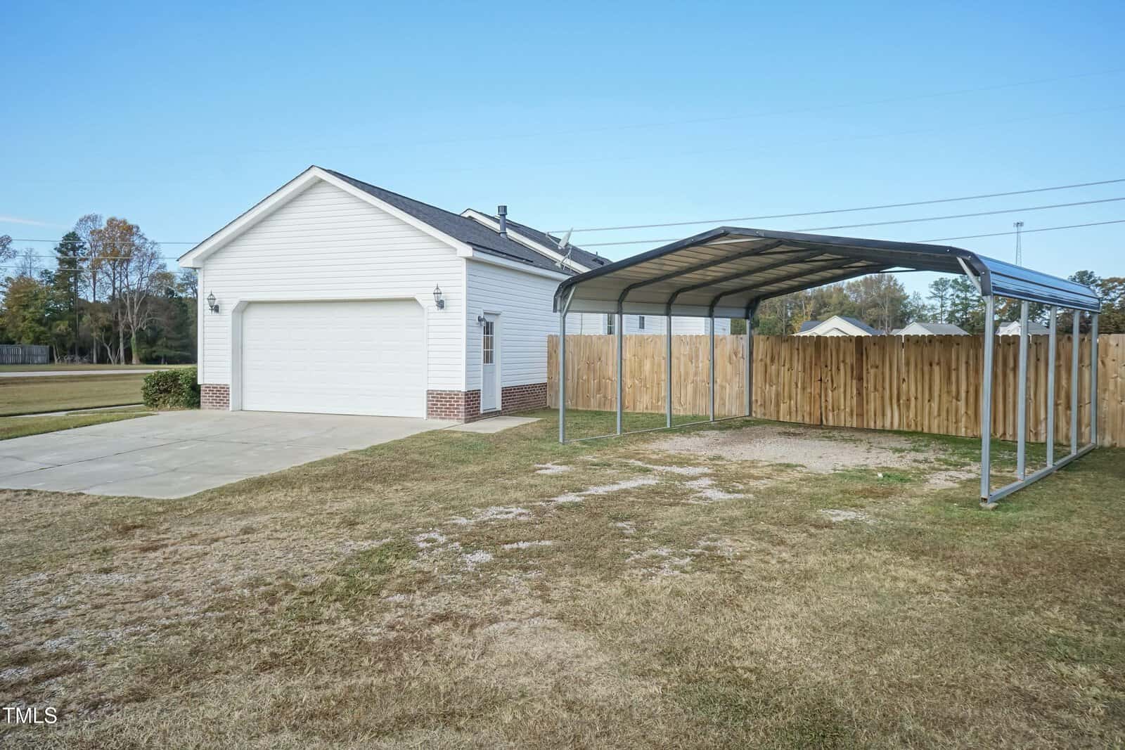 A detached white garage with brick trim, a concrete driveway, and a metal carport beside a wooden privacy fence, set on a large grassy yard under a clear blue sky.