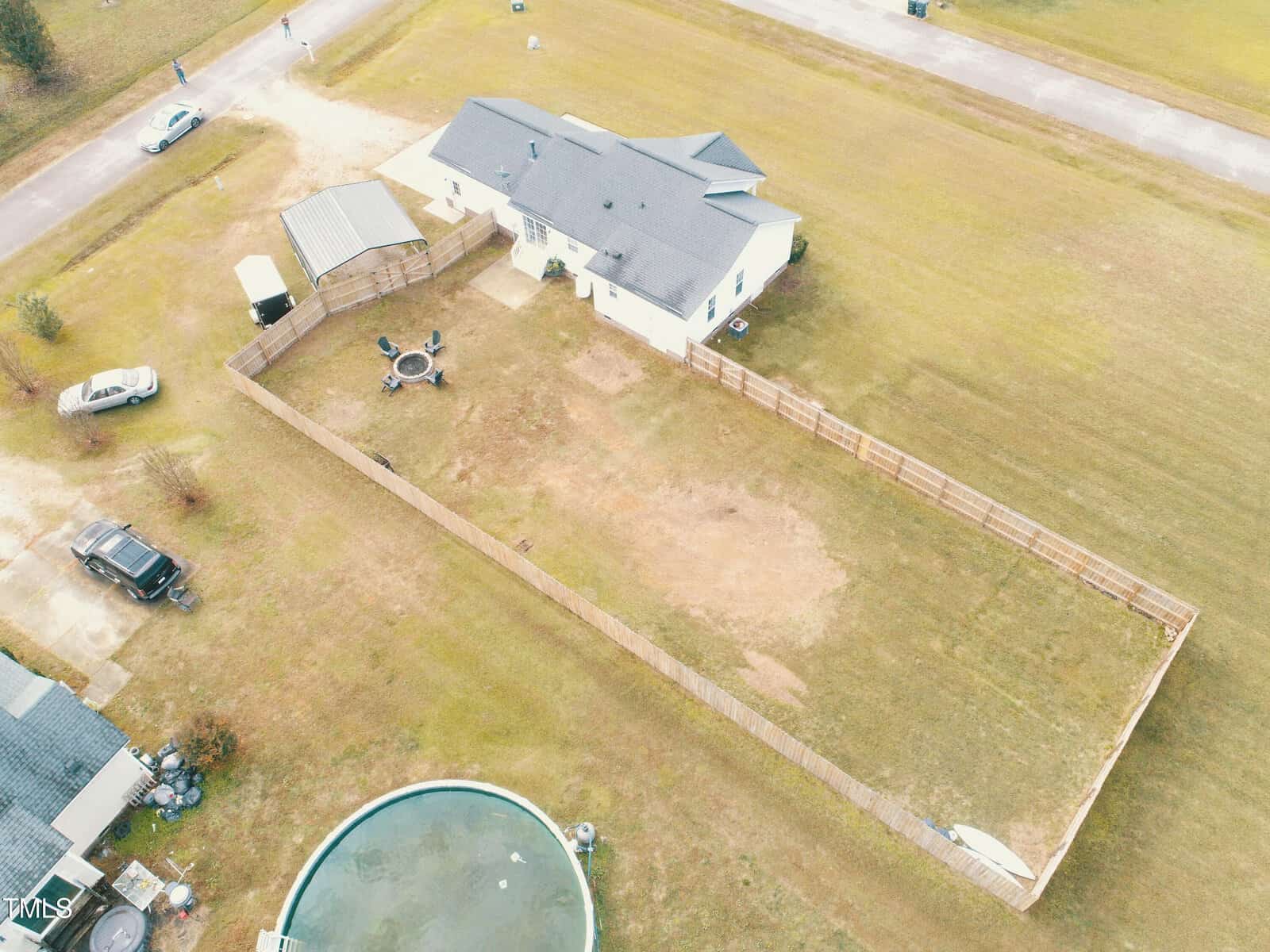 Aerial view of a house with a fenced backyard, containing a fire pit with chairs and a small shed. A round pool is visible outside the fence, and cars are parked nearby on a large grassy lot.