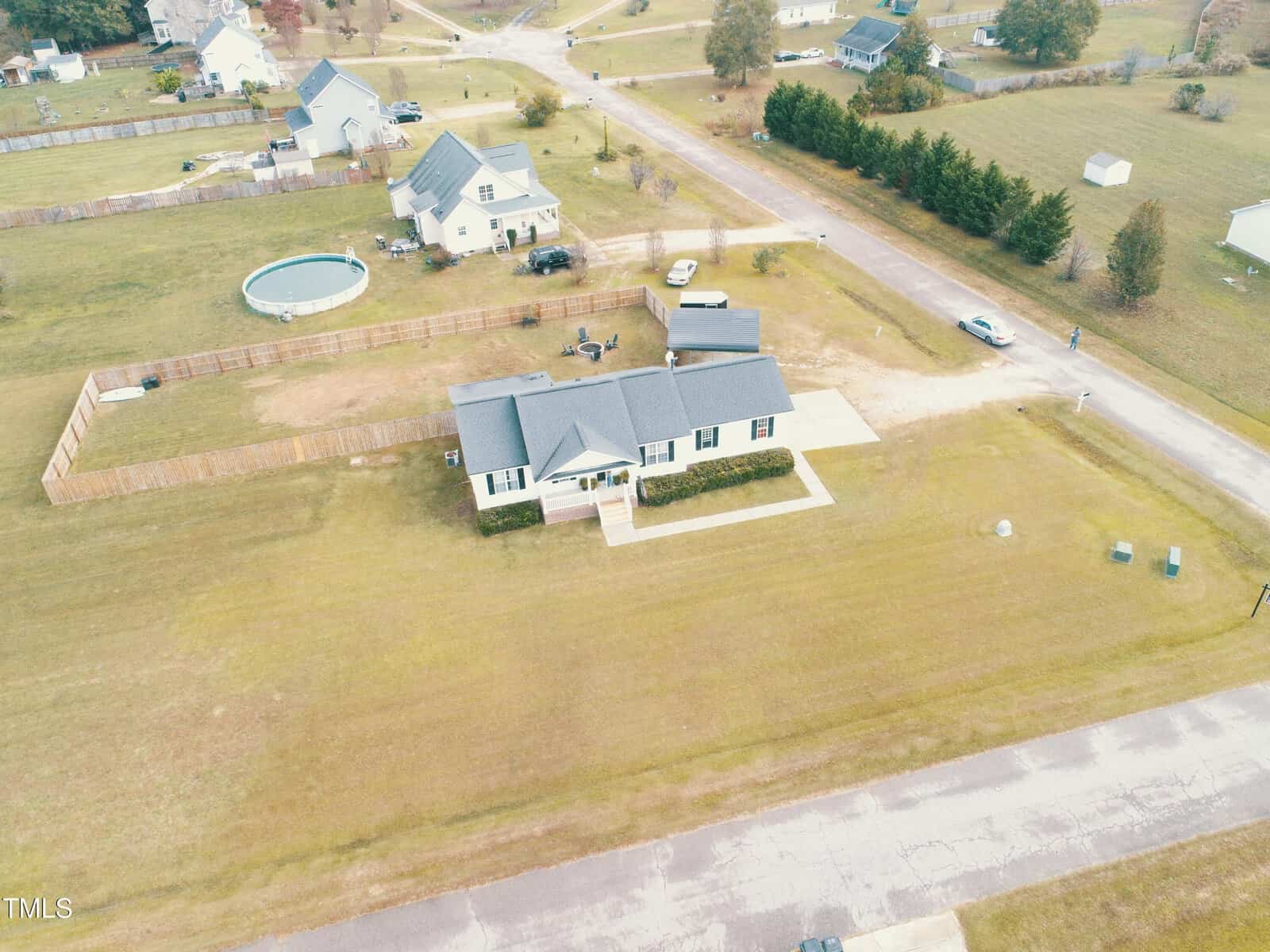 Aerial view of a single-story house with a fenced backyard, a circular above-ground pool, and a shed. The house sits on a large grassy lot in a residential neighborhood with several nearby homes and roads.