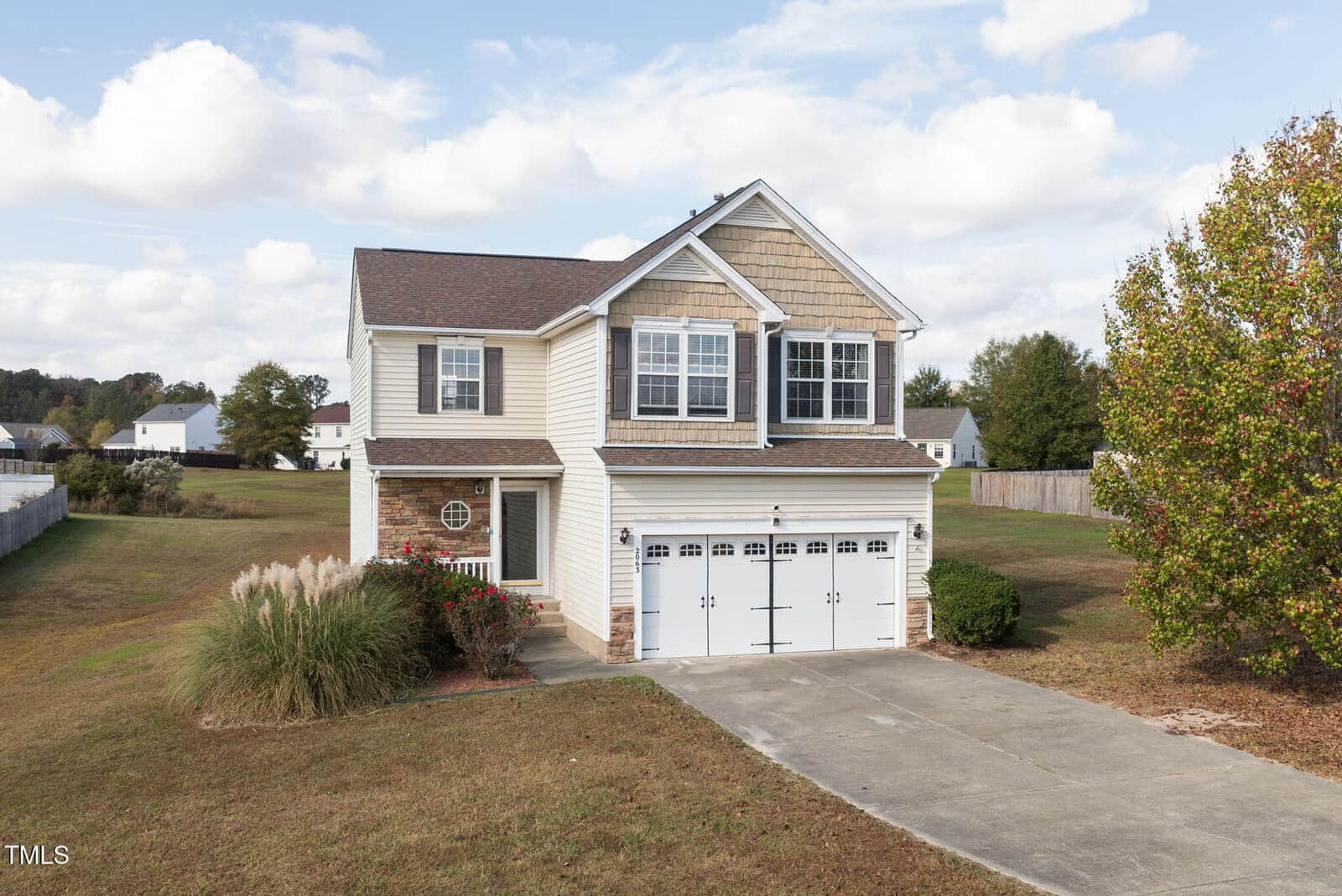 A two-story house with beige siding and stone accent, featuring a double garage with white doors and decorative windows. It's surrounded by a spacious yard with grass and a tree on the right. A cloudy sky is in the background.