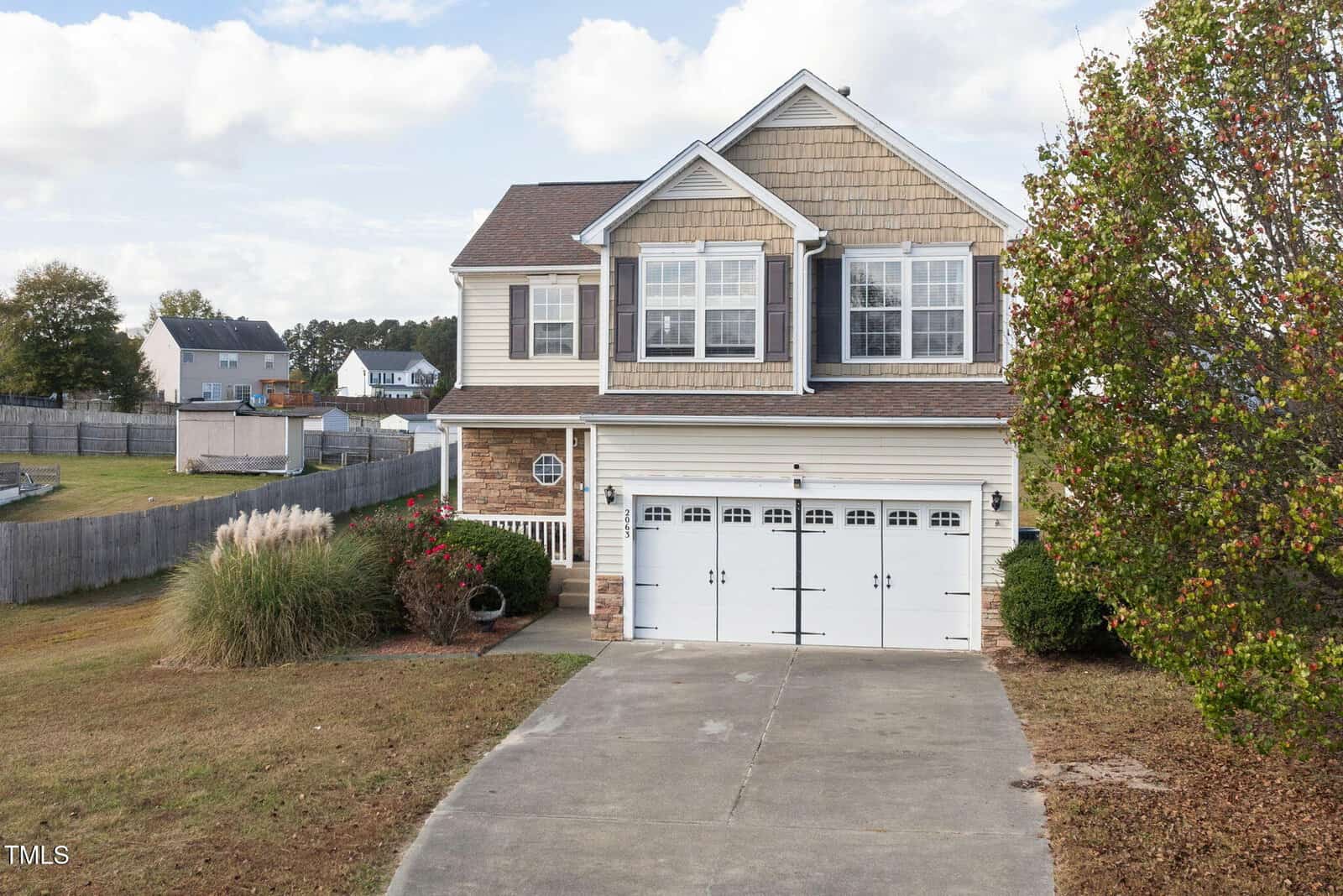 Two-story house with beige siding and brick accents, featuring a double garage door. A concrete driveway leads to the entrance. The yard is landscaped with grass and bushes, and a tree is on the right. Other houses are visible in the background under a cloudy sky.