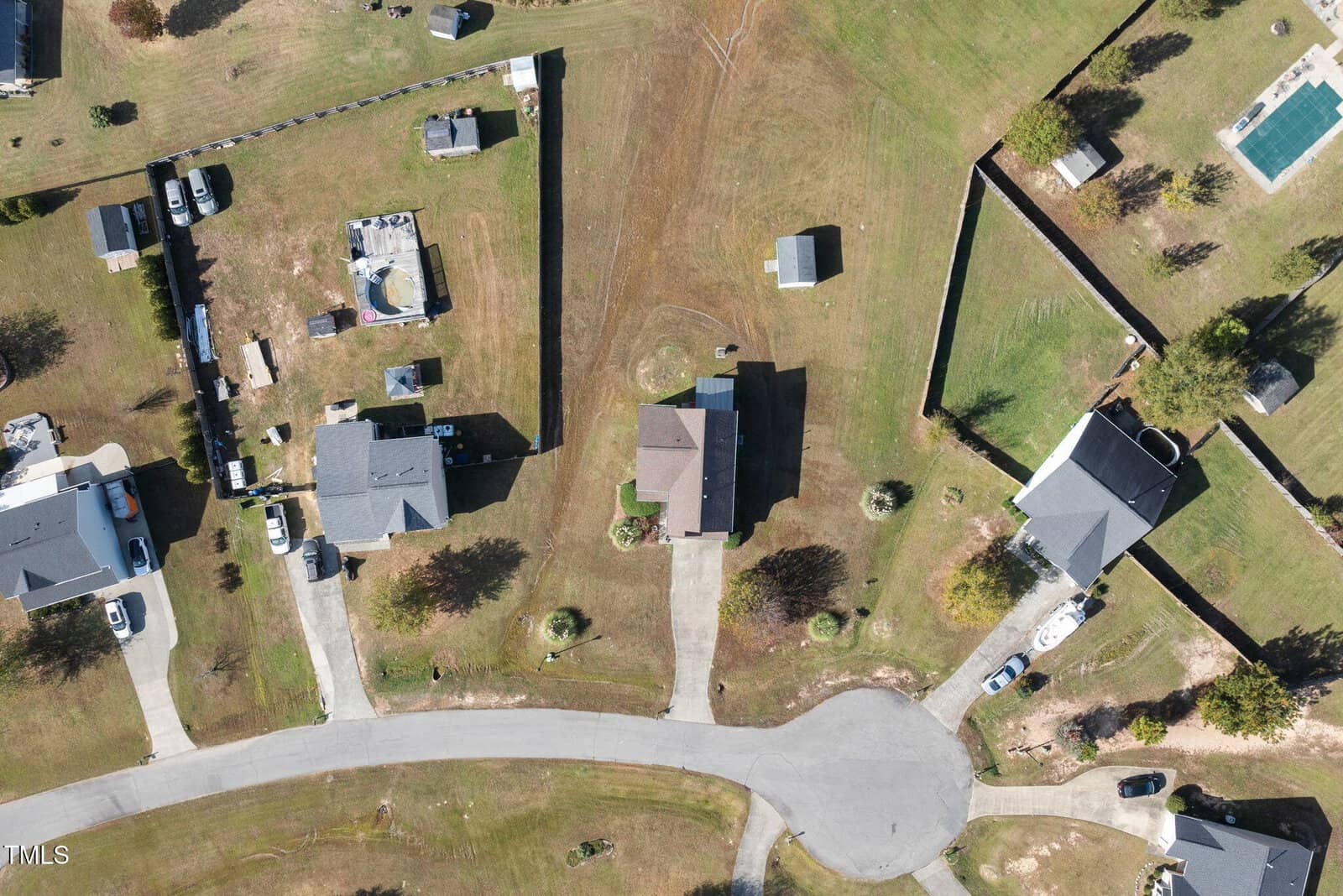Aerial view of a suburban neighborhood showing several houses with driveways, yards, and trees. The image includes a curved road and a few fenced areas, with scattered small structures and a swimming pool.