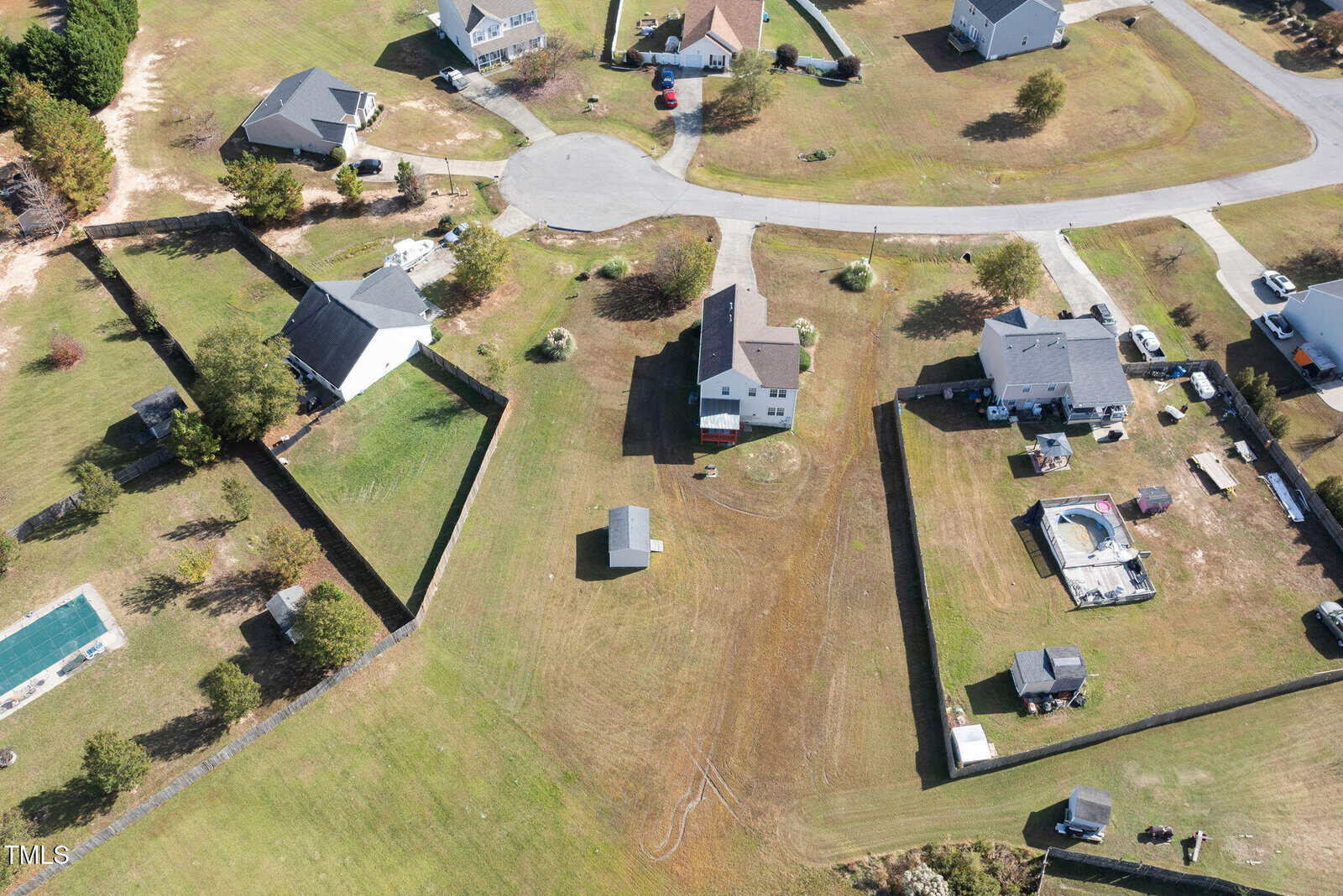 Aerial view of a suburban neighborhood with several houses on large plots of land. The area features fenced backyards, a cul-de-sac, and open green spaces. Some properties have outbuildings and above-ground pools.