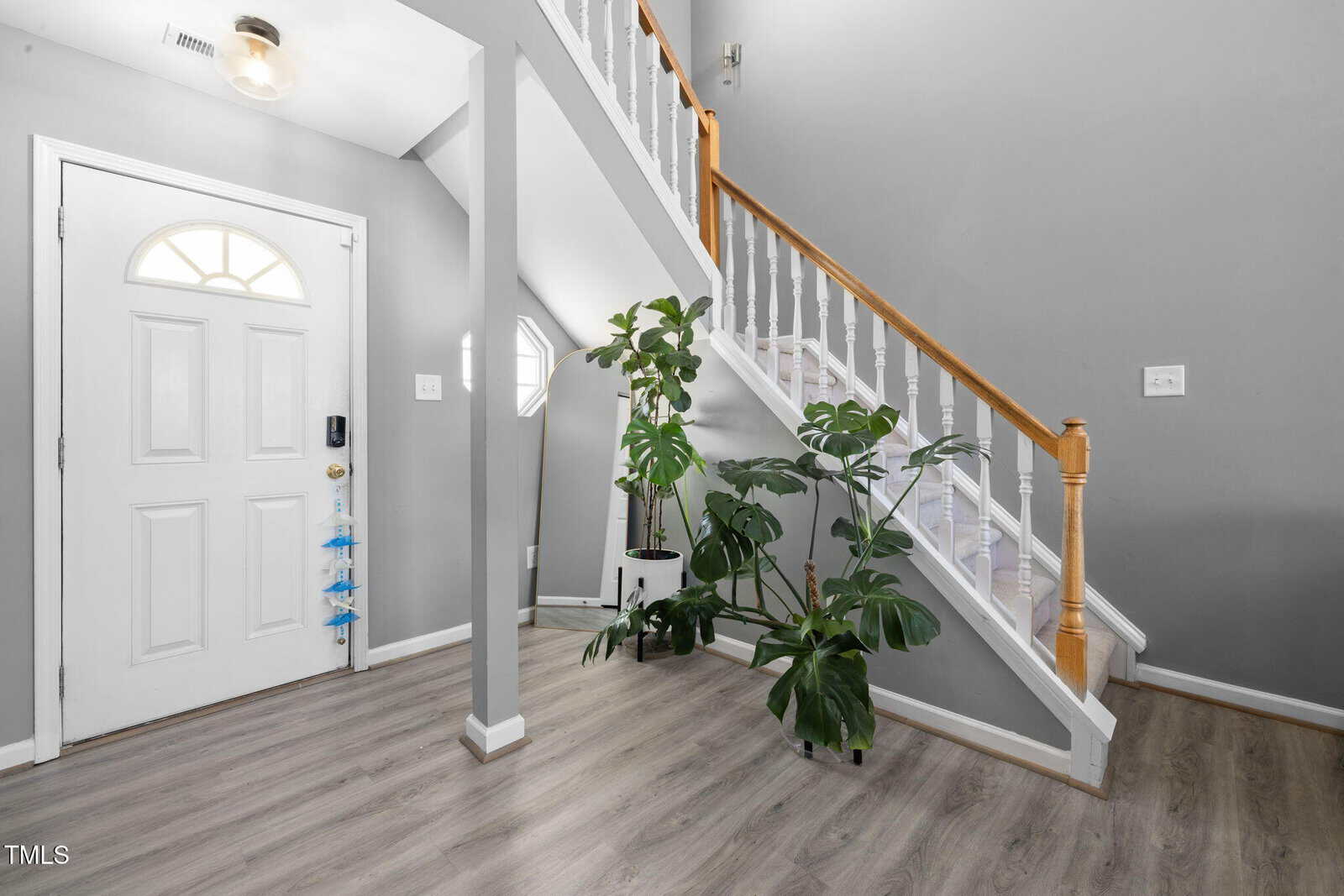 A modern home entryway featuring a light gray color scheme. A small staircase with white railings and wooden accents leads to the upper floor. Potted plants are placed at the base of the stairs, and a white door with a semi-circular window is on the left.