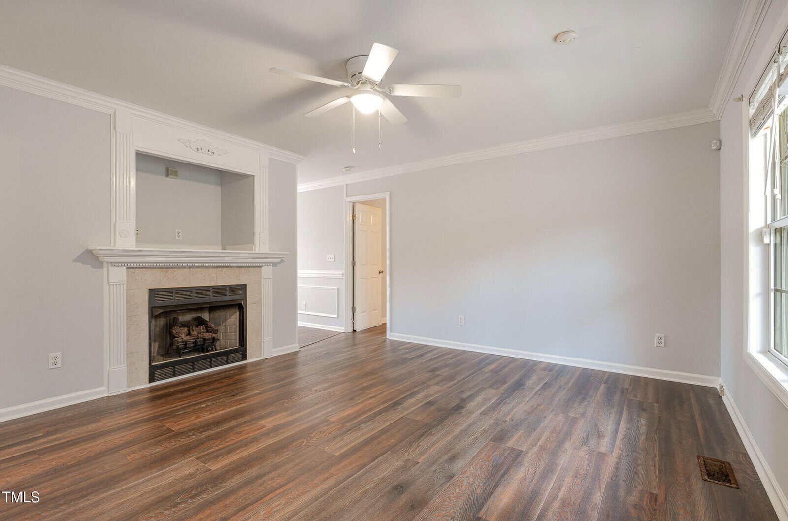 A spacious, empty living room with light gray walls, dark wood flooring, a white ceiling fan, a white fireplace, and a large window letting in natural light.