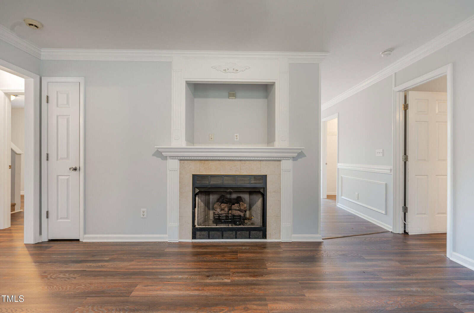 A living room with hardwood floors, light gray walls, a white fireplace with a mantel, and a built-in alcove above the fireplace. There are two doors and a hallway leading to other rooms.
