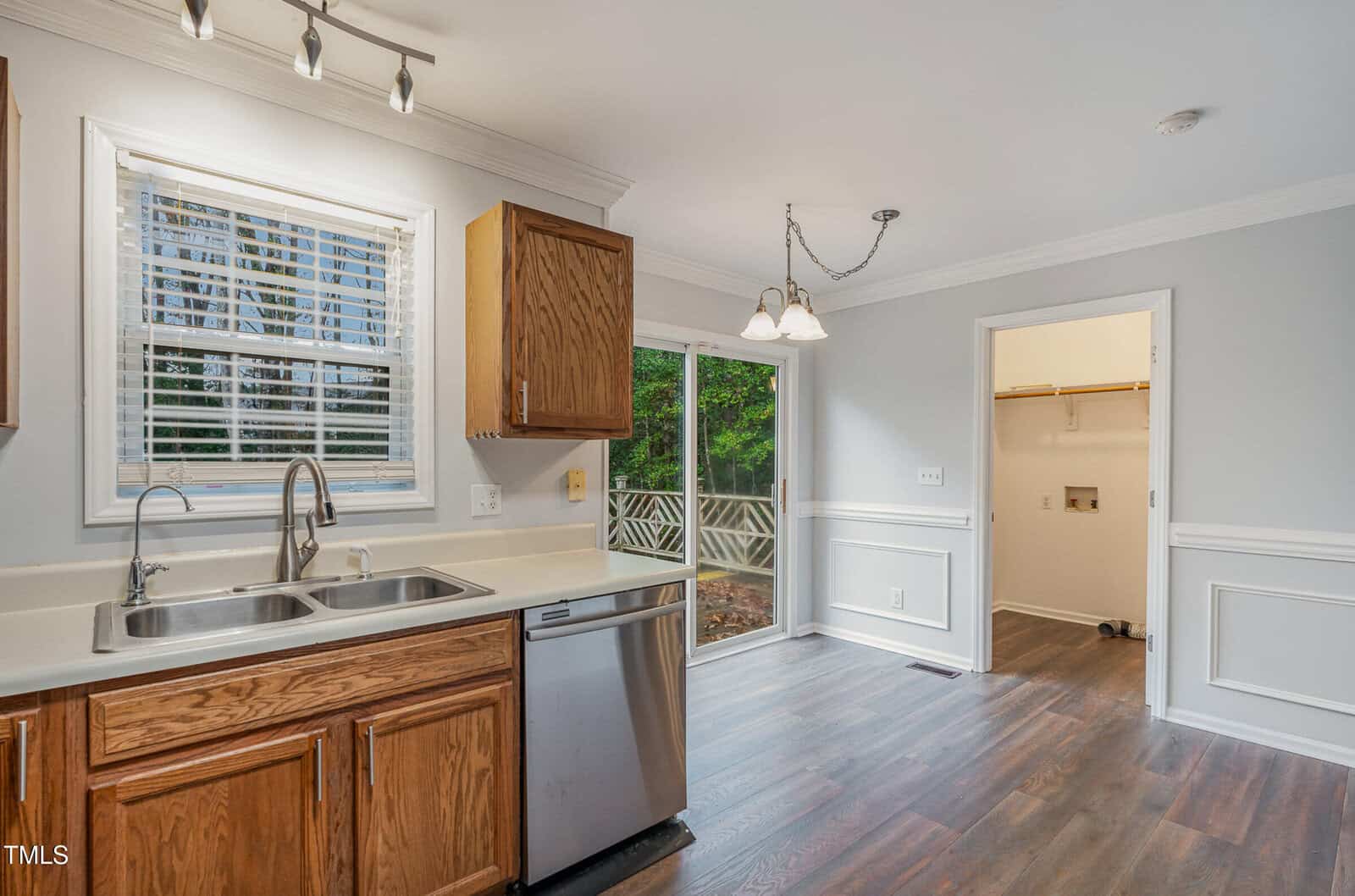 Kitchen with double sink, stainless steel dishwasher, and wooden cabinets. A window is above the sink, and sliding glass doors lead to a deck. Adjacent room contains a laundry area with hookups. Dark wood flooring throughout.