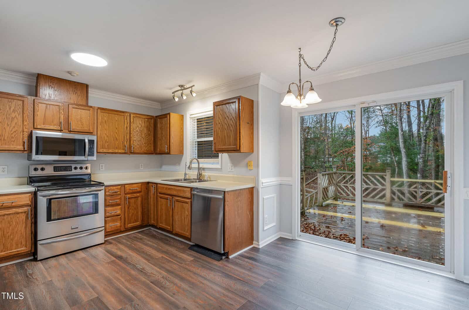 Modern kitchen with stainless steel appliances, wooden cabinets, and dark wood flooring. A sliding glass door leads to a deck outside with trees visible in the background. Light fixture hangs above the dining area.