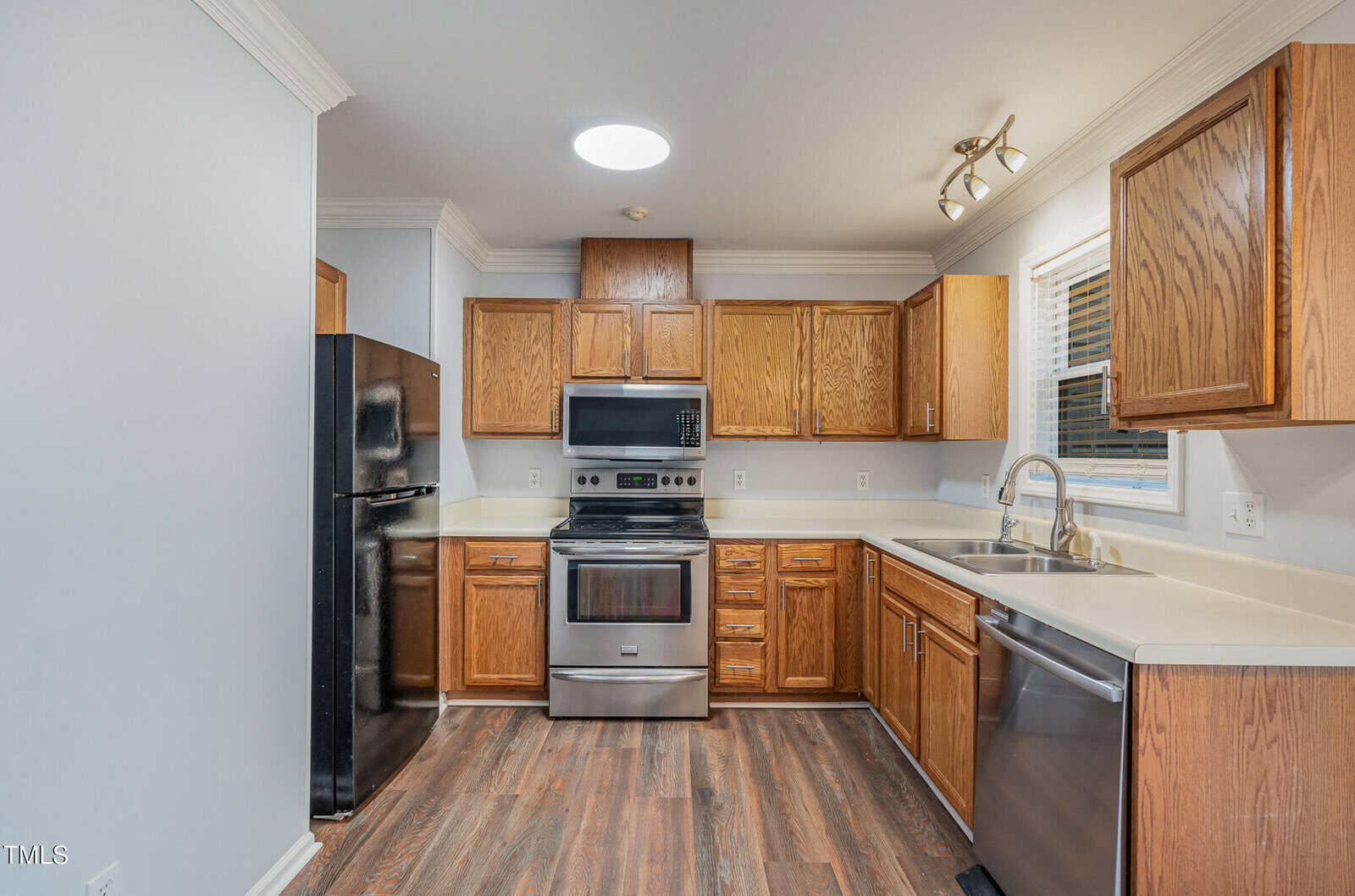 Modern kitchen with wooden cabinets, stainless steel oven and dishwasher, black refrigerator, electric stove, under-cabinet lighting, and a single window above the sink. Wood flooring and light-colored walls.