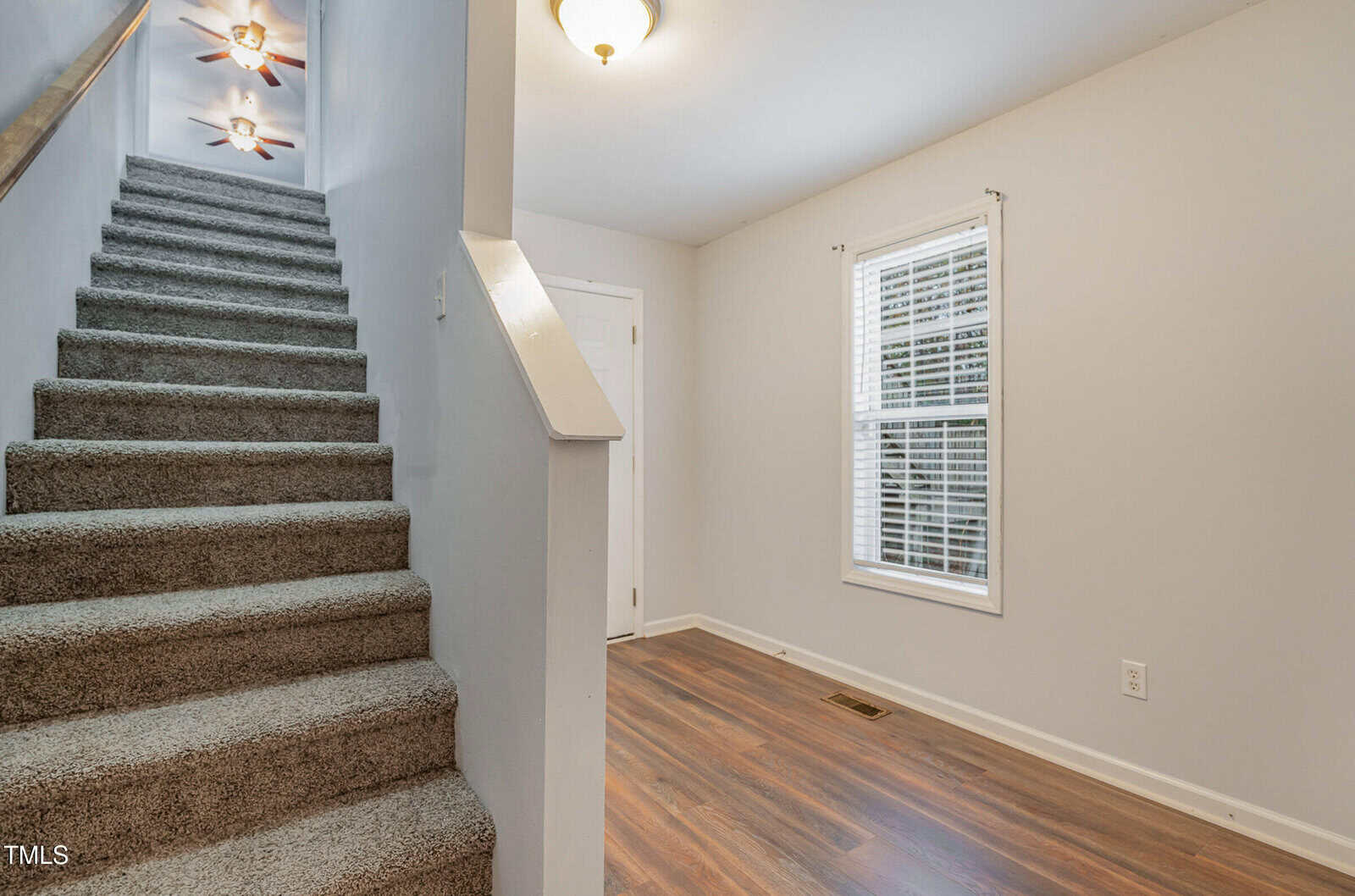 Carpeted stairs with a white railing lead up from a hallway with wood flooring, white walls, a window with blinds, and a ceiling light fixture.