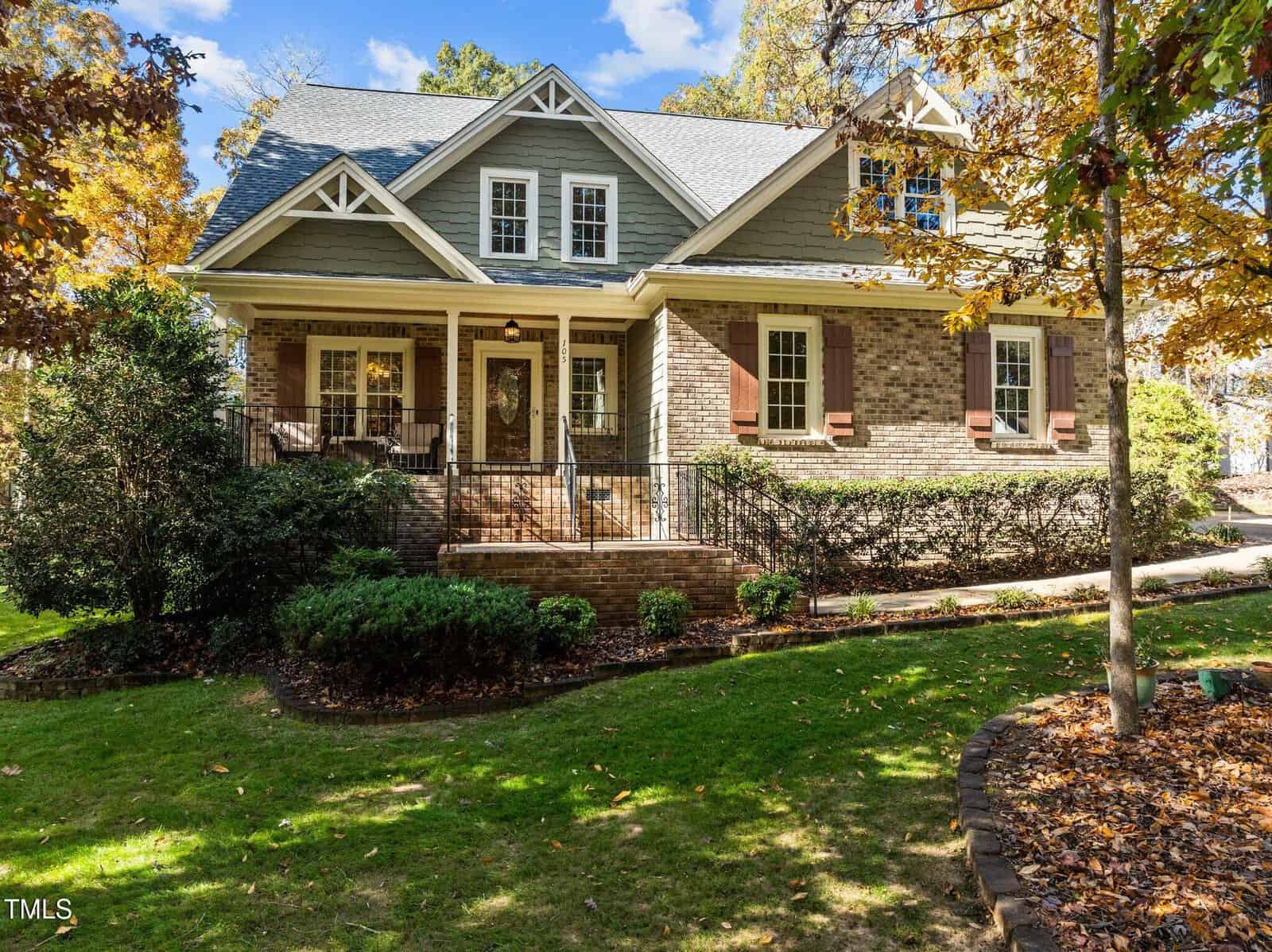 A two-story house with gray siding and brick exterior, white trim, brown shutters, and a covered front porch with black railing, surrounded by green lawn, bushes, and trees with autumn leaves.