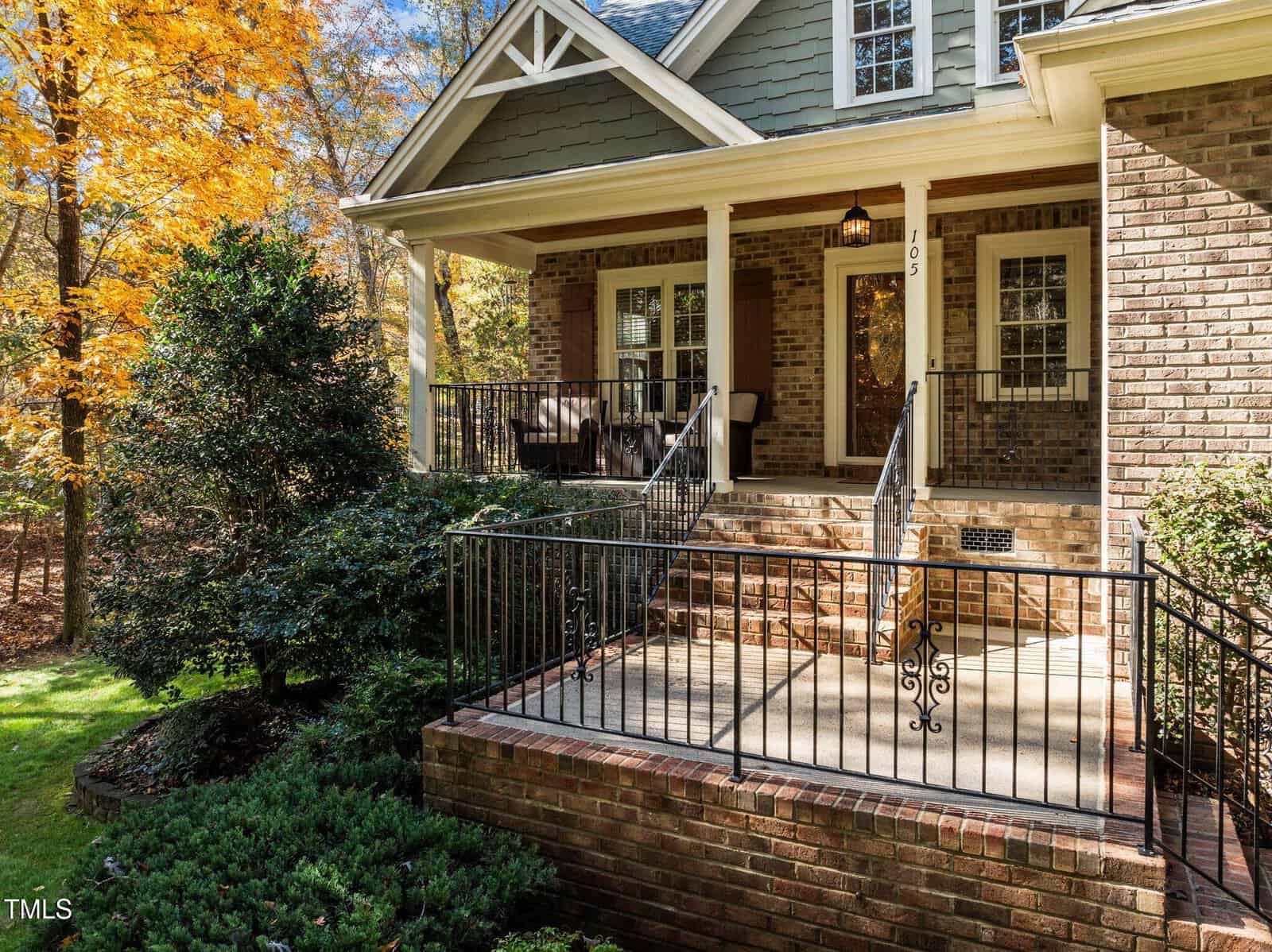 A brick house with a covered front porch, black metal railings, and steps leading up to the entrance. There are chairs on the porch, bushes in front, and trees with autumn leaves in the background.