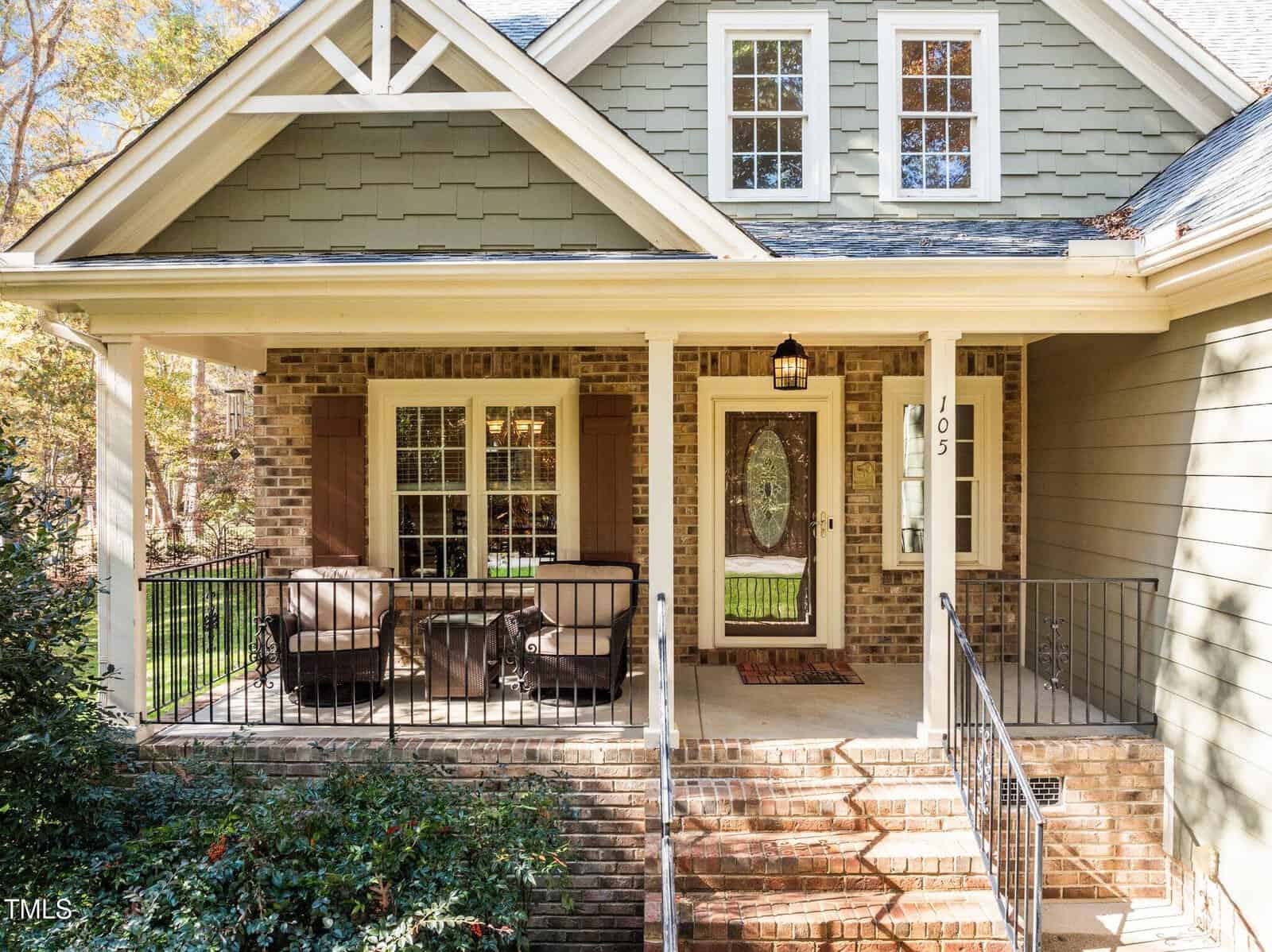 A front porch of a house with brick and green siding, featuring a seating area with two cushioned chairs and a table. There are stairs with black railings leading up to a wooden front door with glass panels.