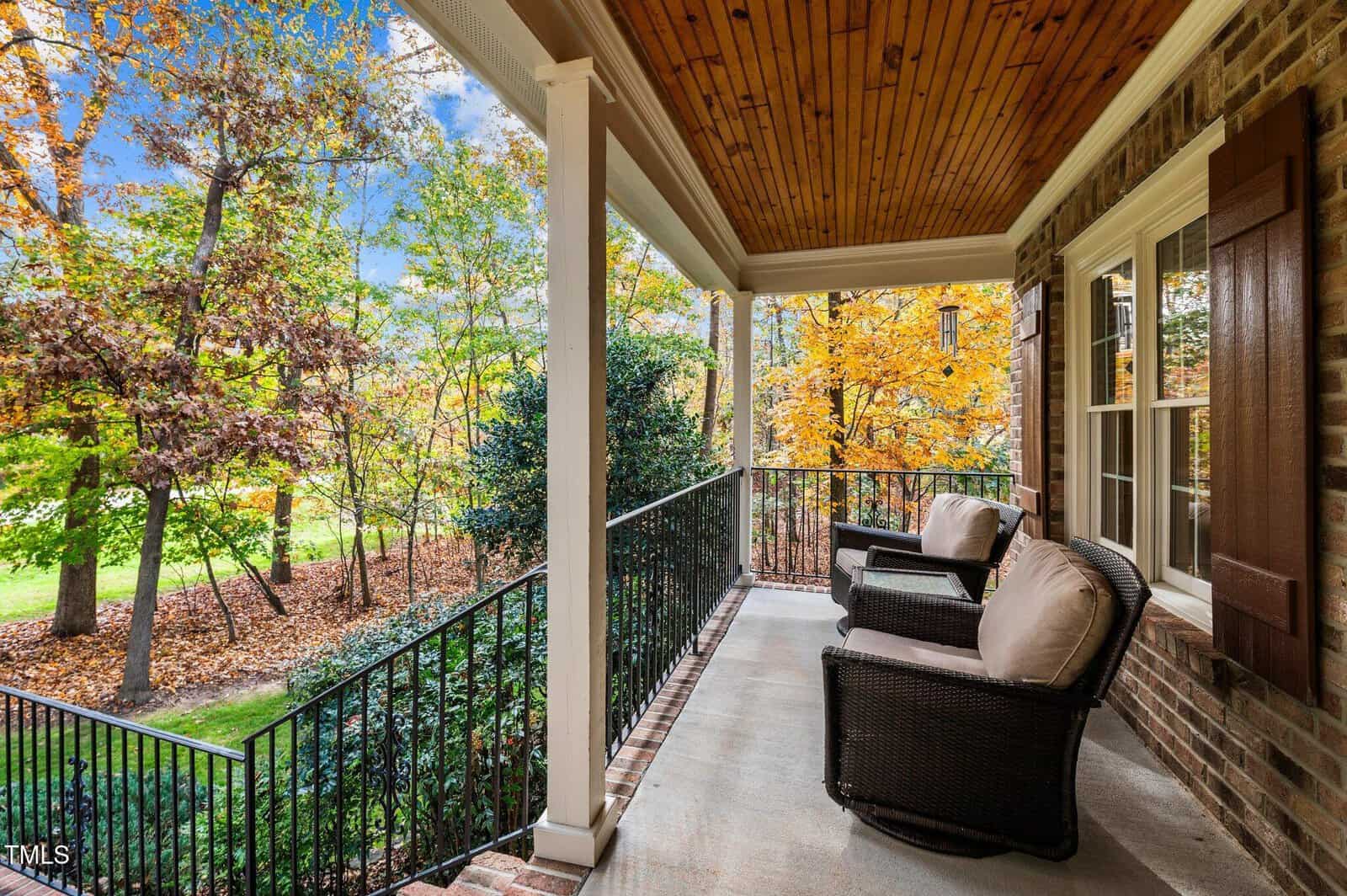 A covered porch with two cushioned wicker chairs overlooks a yard with trees displaying autumn foliage. The porch has a wooden ceiling, black metal railing, and brick exterior walls with windows and shutters.