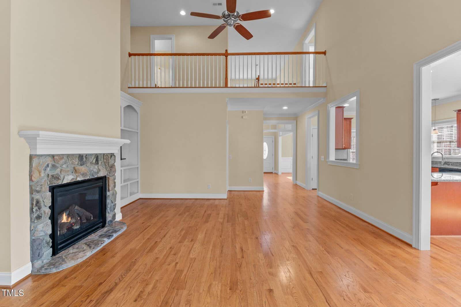 Spacious living room with hardwood flooring, stone fireplace, ceiling fan, upper balcony railing, and open doorways leading to kitchen and other rooms. Neutral beige walls and white trim throughout.