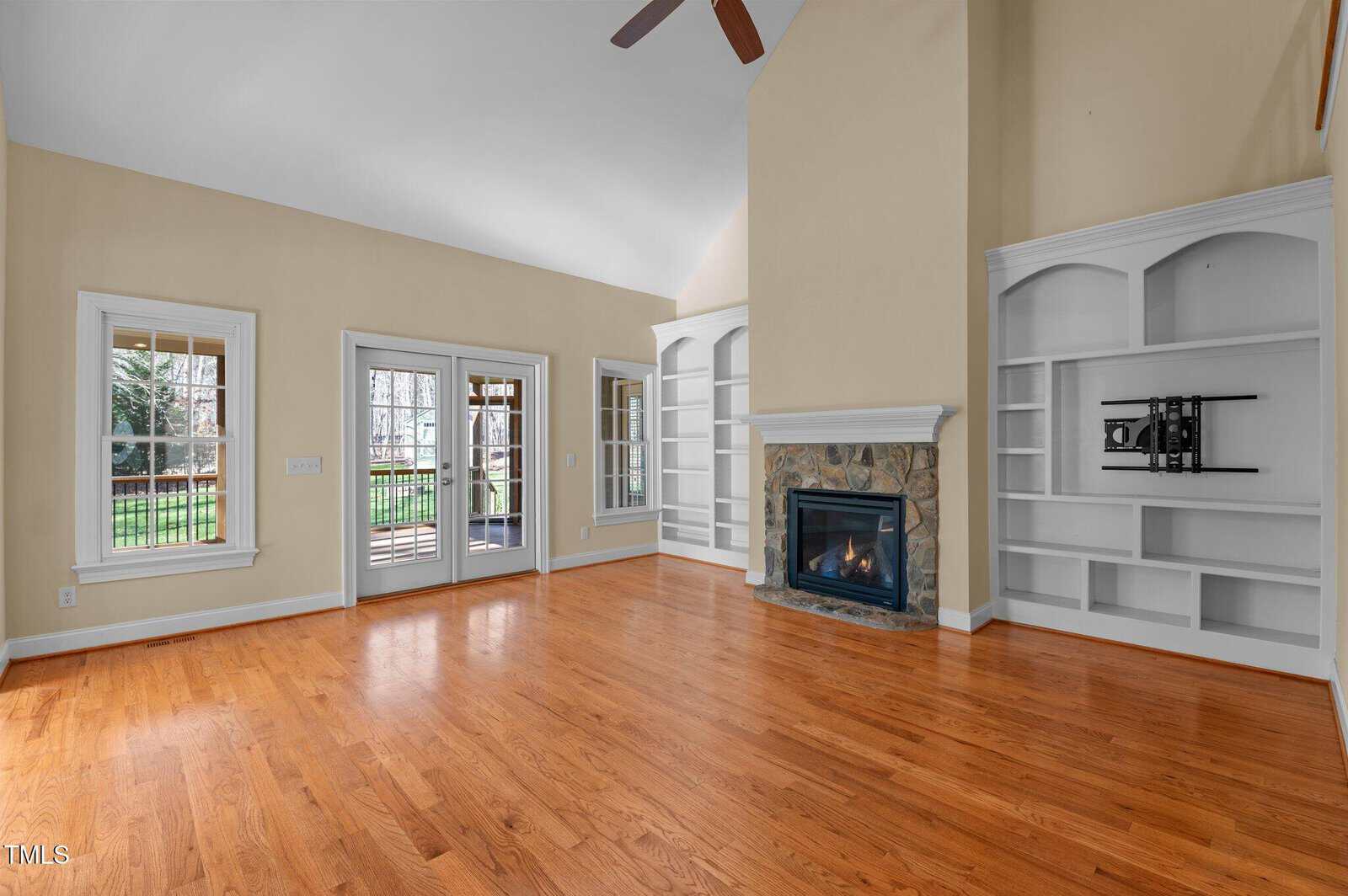 A spacious living room with hardwood floors, built-in white shelves, a stone-faced fireplace, large windows, and glass doors leading to a fenced backyard. The walls are light beige and the ceiling is vaulted.