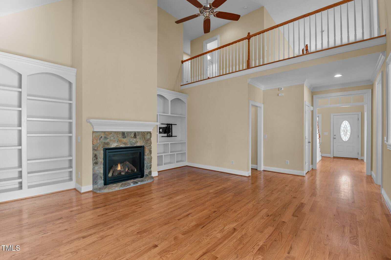 Spacious living room with hardwood floors, built-in white shelves, a stone fireplace, high ceiling with fan, and an upstairs balcony. A hallway leads to the front door with a decorative glass window.