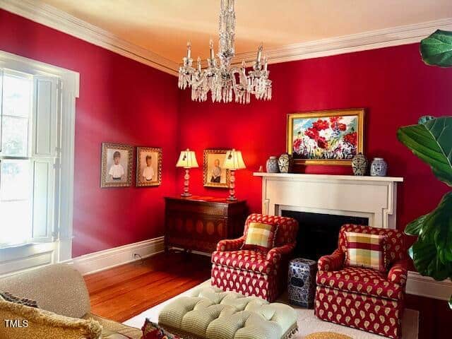 A brightly lit living room with red walls, chandelier, and white trim. Two red patterned chairs face a fireplace adorned with art and vases. A cushioned ottoman sits in front, flanked by a couch and a window allowing natural light.