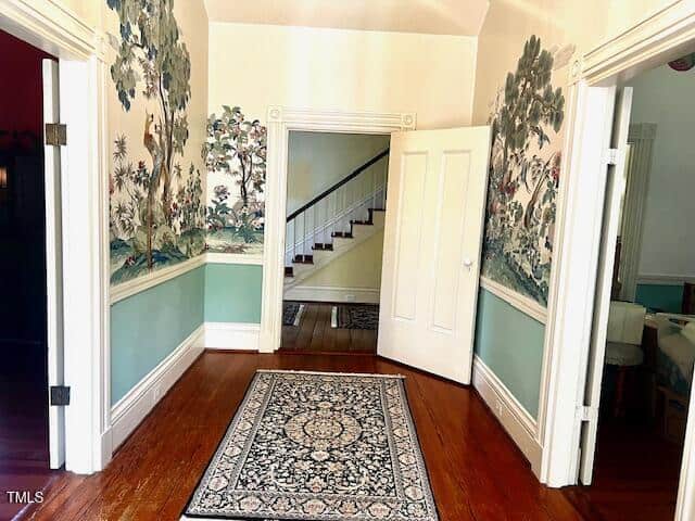 A hallway with wooden floors features floral wallpaper above green wall paneling. A patterned rug lies on the floor. An open doorway reveals stairs with a white railing and green-painted walls. Additional open doors lead to other rooms.
