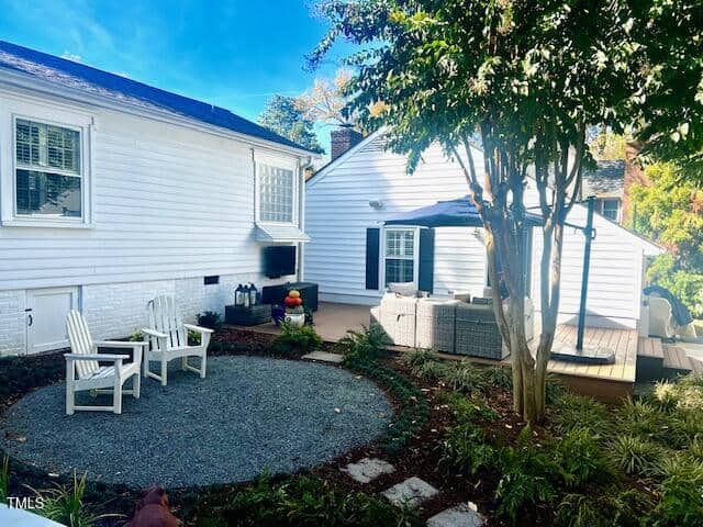 A backyard featuring a circular gravel area with white Adirondack chairs surrounding a small fire pit. A deck with outdoor furniture and an umbrella is adjacent, under a tall tree. The house is white with several windows, and the sky is clear and blue.
