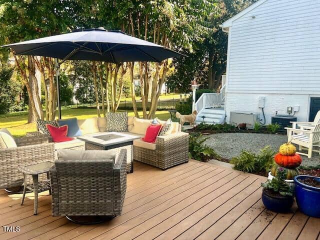 A cozy wooden deck features a wicker outdoor seating set with cushions, a central fire pit, and a large umbrella. A dog stands nearby. Potted plants and a stack of pumpkins add decoration. Trees and a white house frame the background.