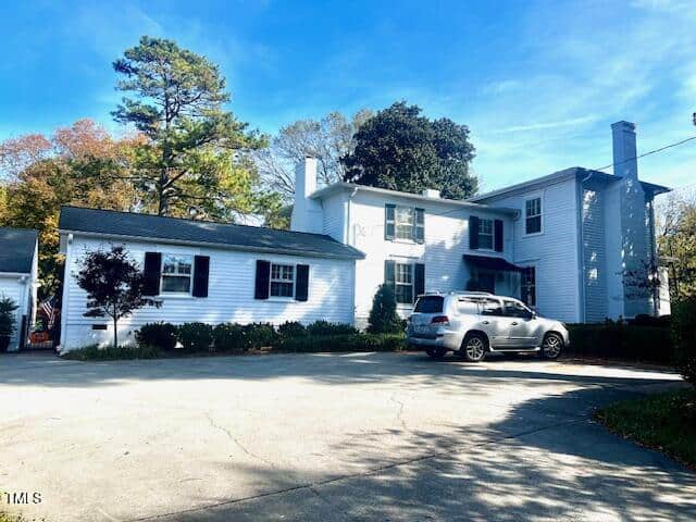 A two-story white house with black shutters and a gray roof, surrounded by trees. A silver SUV is parked in the driveway under a clear blue sky.