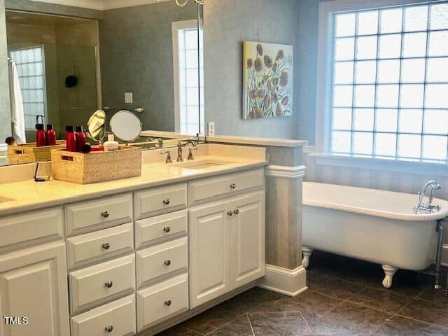 Elegant bathroom with a clawfoot tub near a large frosted window. A double-sink vanity with white cabinets is topped with baskets and toiletries. Walls feature subtle patterned wallpaper and a floral painting.