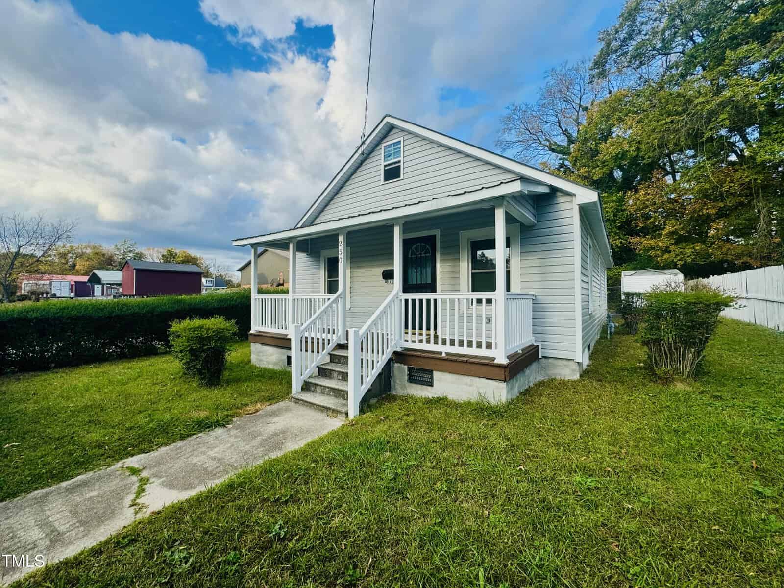A small gray house with a white porch and steps, set in a grassy yard. The sky is partly cloudy, with a few trees and a red building in the background.
