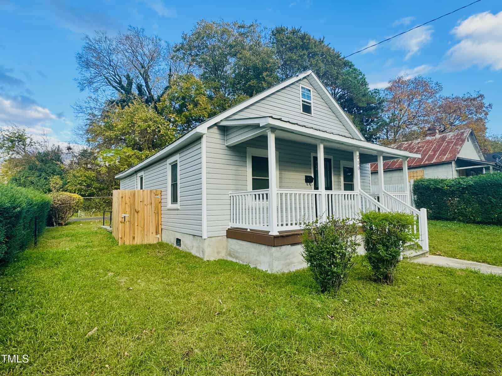 A small, light gray house with a white porch railing sits surrounded by green grass and trees. A wooden fence is visible to the left, and neighboring houses can be seen. The sky is partly cloudy, adding a serene atmosphere to the scene.