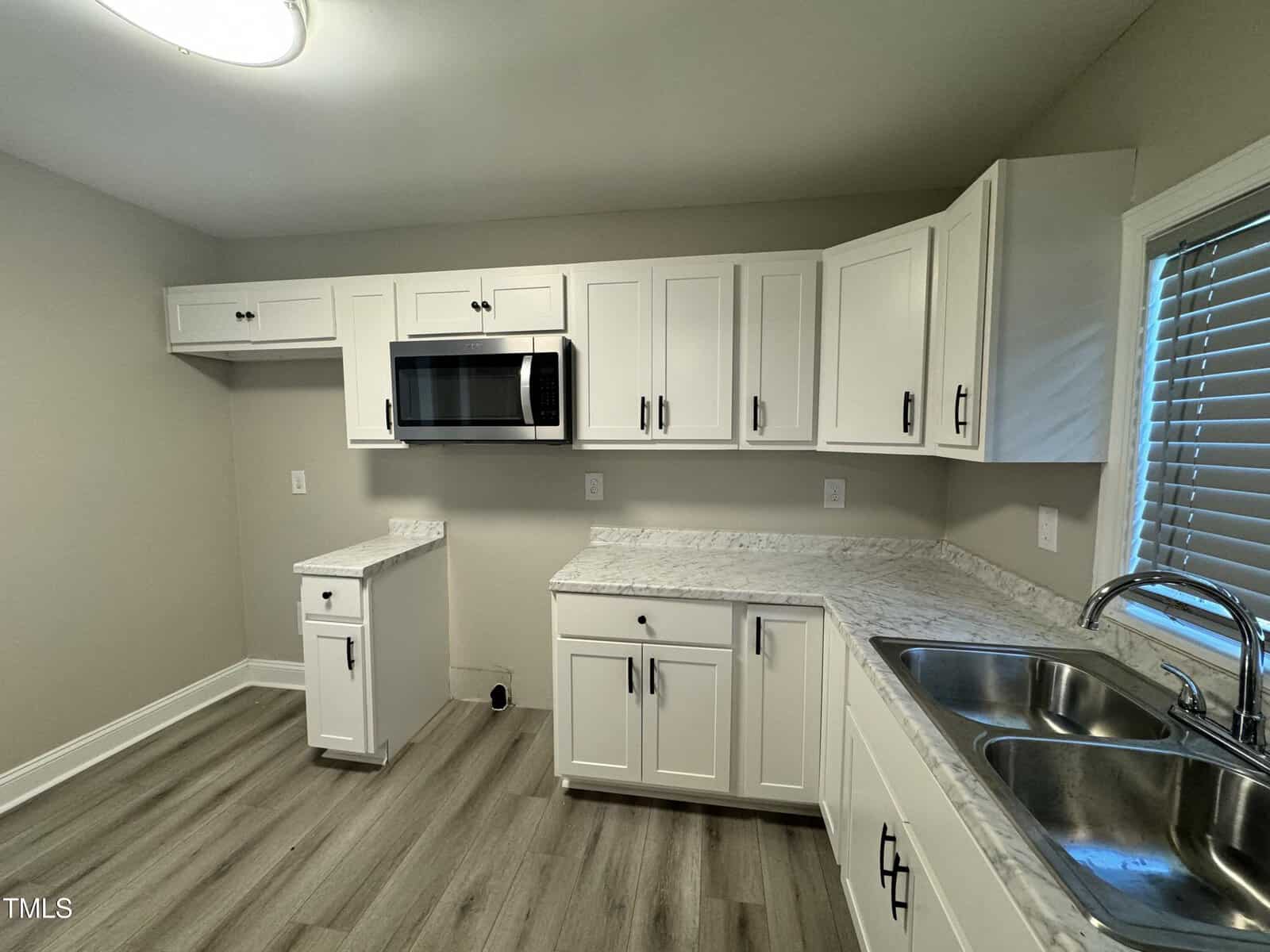 A modern kitchen with white cabinets, a stainless steel microwave, and a dual sink. The countertops are marble, and the floor is wood. A window with blinds is on the right, allowing natural light to enter the space.