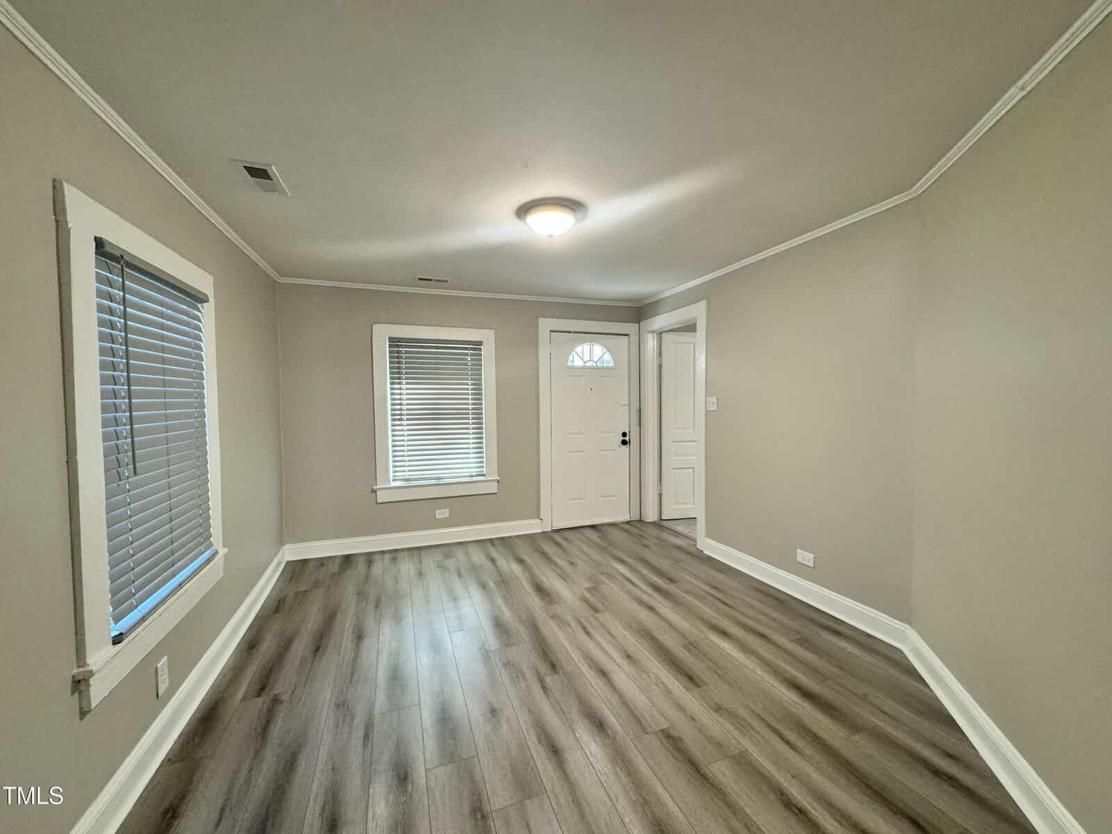 A vacant room with light wood laminate flooring and beige walls. It features two windows with blinds on the left and a white front door with an arched window. A round ceiling light fixture is centrally located.