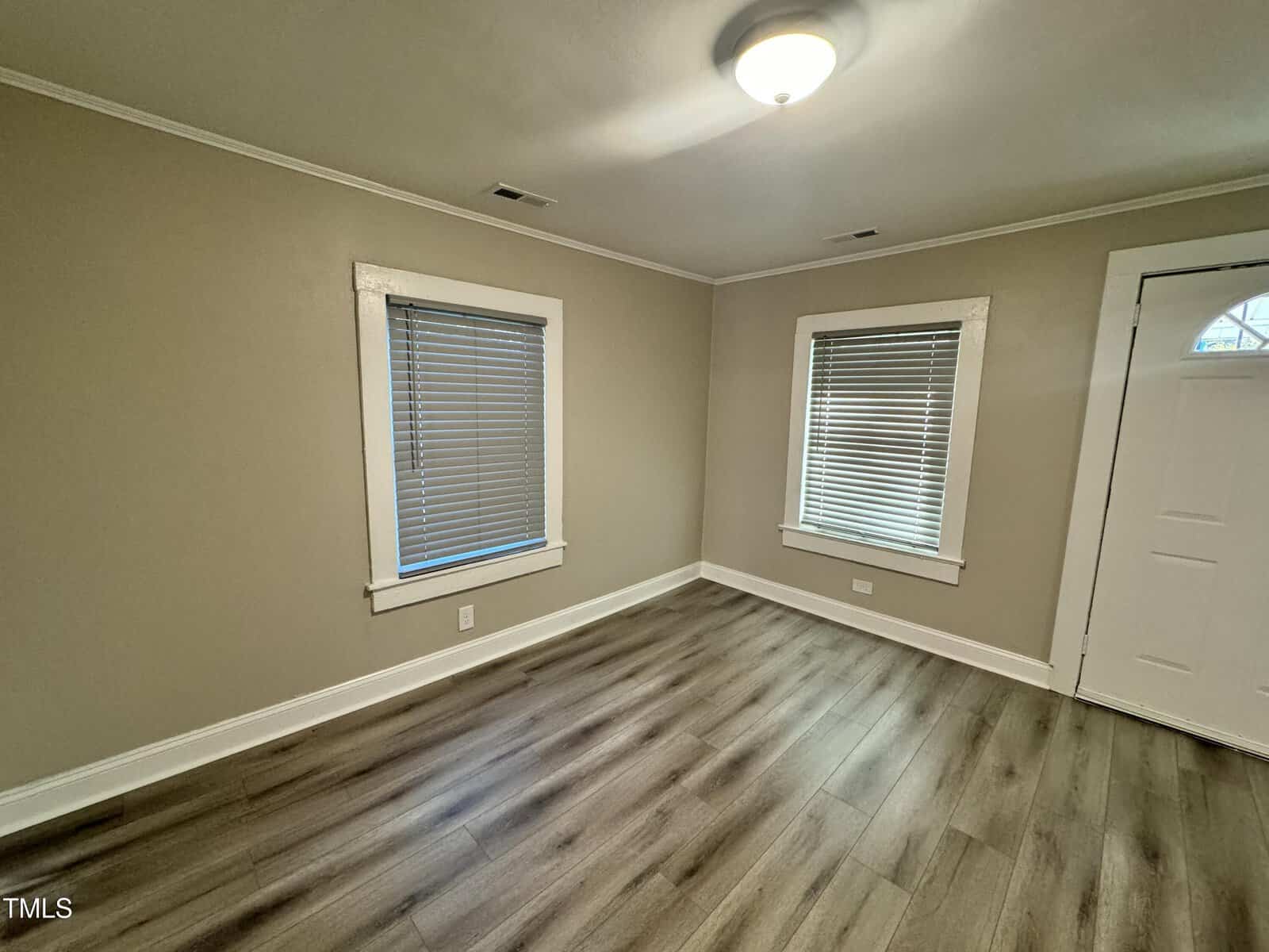 A small room with light gray walls and a wood-patterned floor. Two windows with closed blinds are on the left wall, and a white door on the right. A ceiling light illuminates the space. Trim is white, and two vent openings are visible.