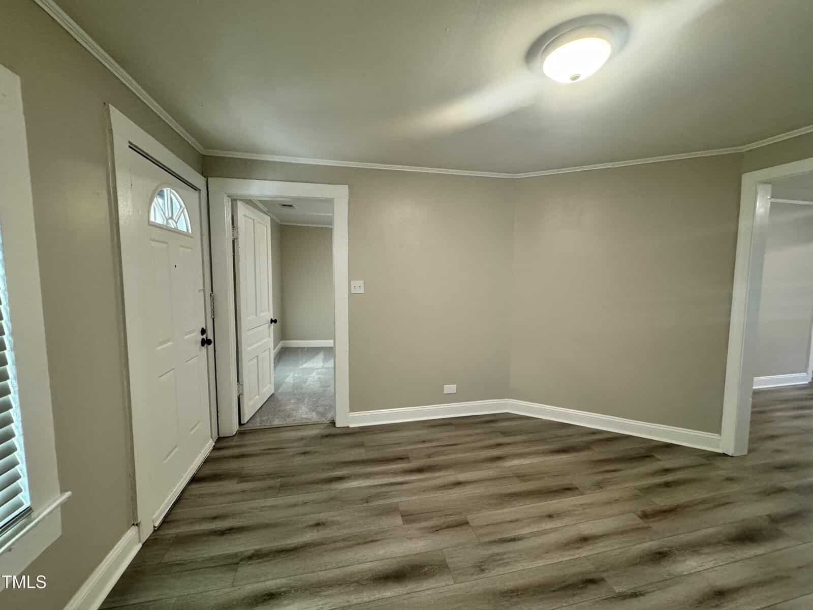 Interior of a room with light-colored walls and wood-patterned flooring. A closed white door with window is on the left, leading to a hallway. A ceiling light fixture is on, and there's a large doorway on the right.