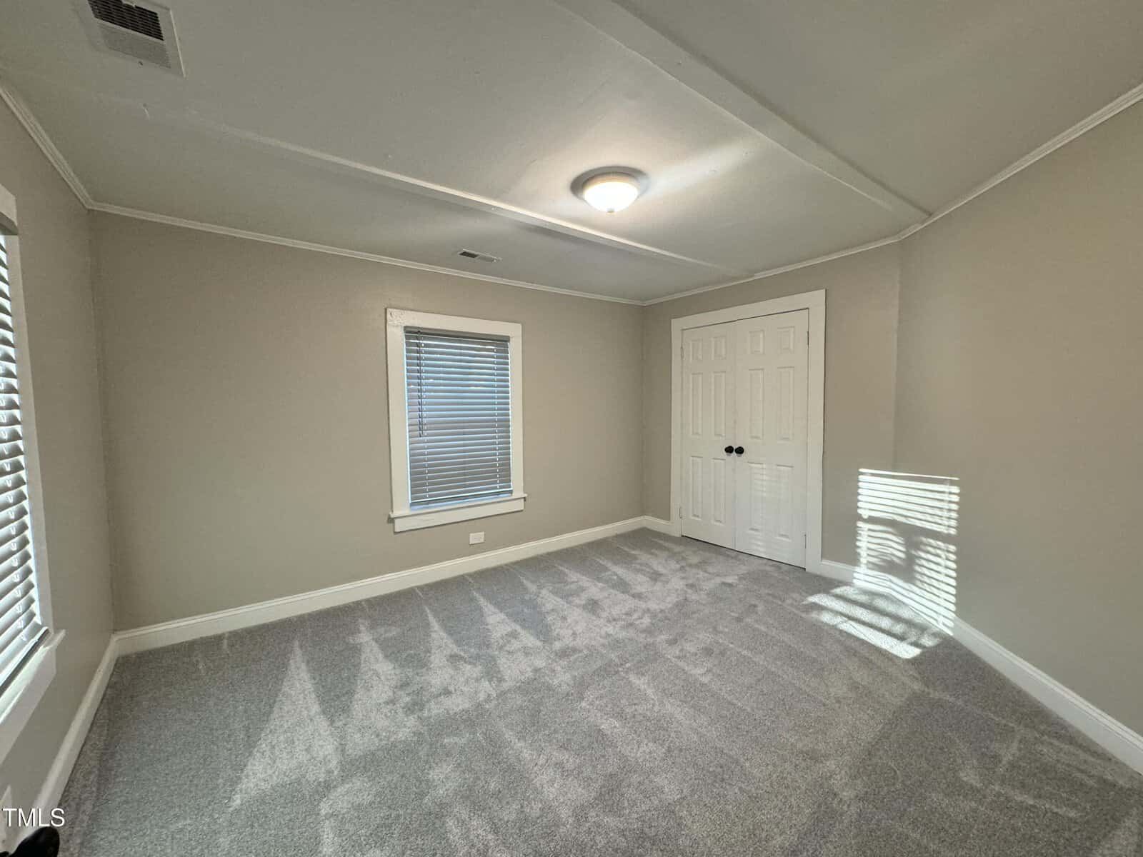Empty room with beige walls, a carpeted floor, and a window with closed blinds. There is a white ceiling light and a closed double door closet. Sunlight partially illuminates the room, creating shadows on the floor.