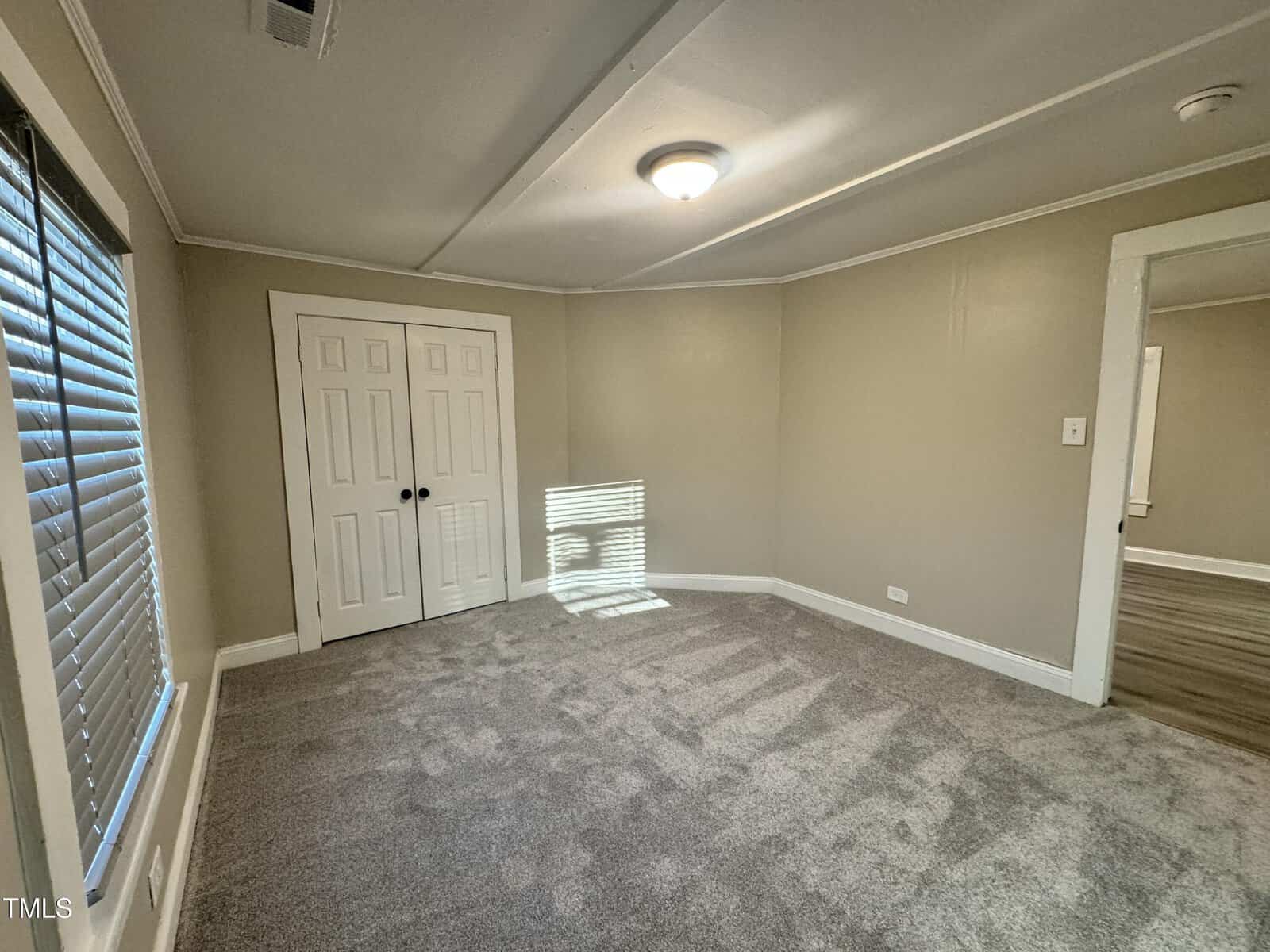 A small, empty room with beige walls and gray carpet. A double white closet door is on one wall, and a window with blinds lets in natural light, casting shadows on the floor. A ceiling light fixture is centered above.