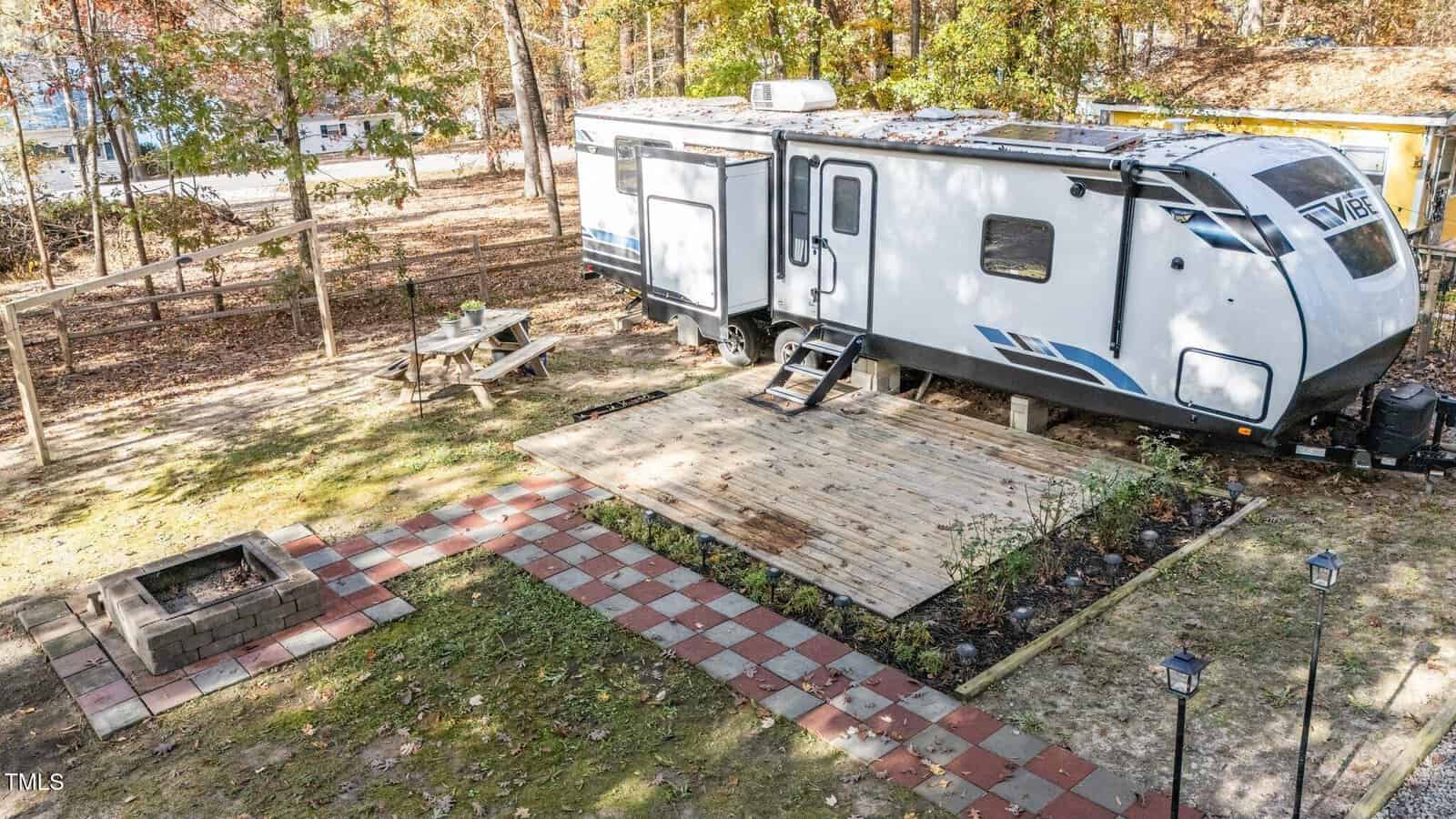 A travel trailer is parked on a grassy lot with a wooden deck, a picnic table, a fire pit built from bricks, and a paved walkway. Trees with autumn leaves surround the area, and a lake is visible in the background.