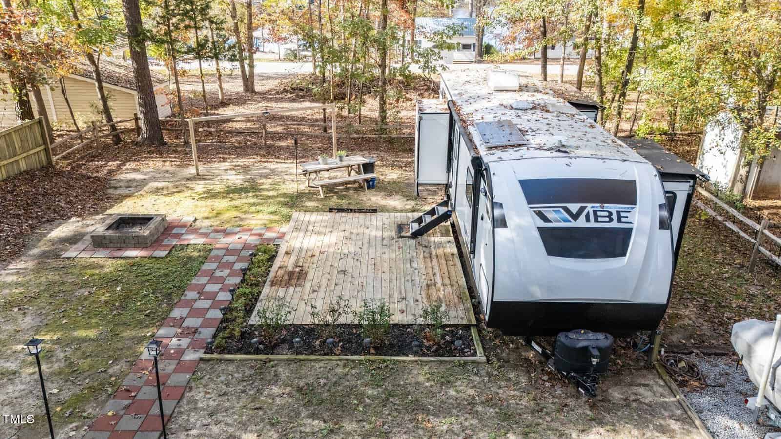 A white Vibe travel trailer is parked on a wooden deck in a fenced backyard with trees. A walkway made of red and white tiles leads to a fire pit area. Leaves are scattered on the trailer and ground. A lake is visible in the background.