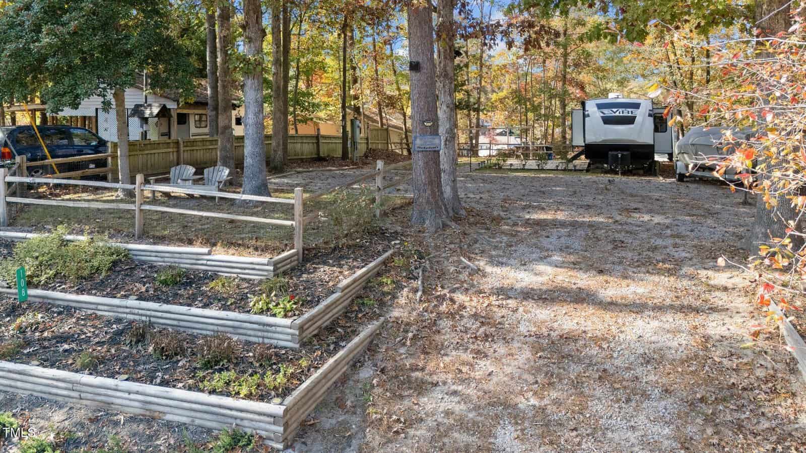 A gravel driveway leads to a parked RV among tall trees. To the left, tiered wooden garden beds hold plants, and two white chairs sit near a wooden fence. Autumn leaves cover the ground.