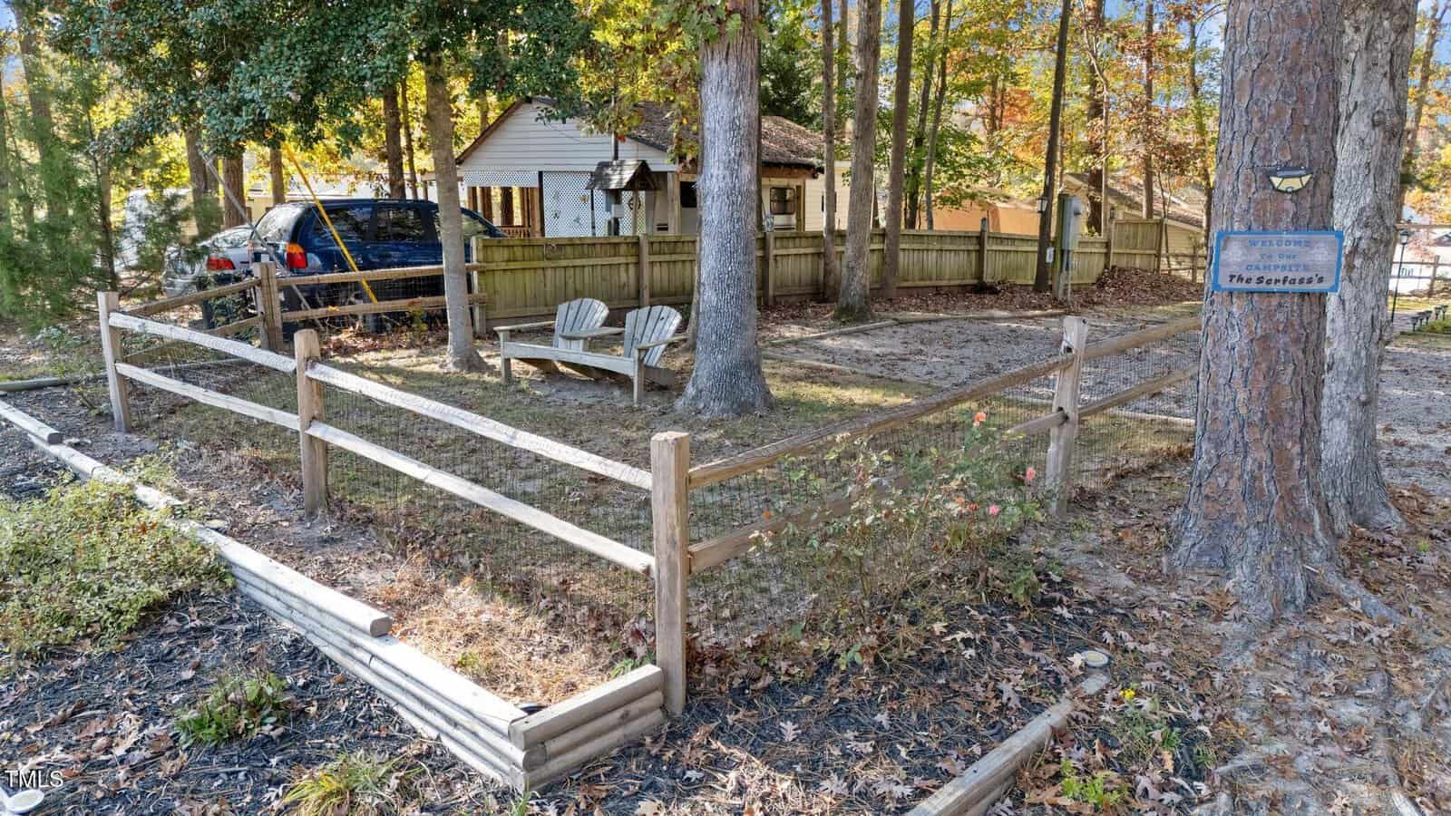 A fenced yard with two wooden chairs under trees, a parked car, and a small house in the background. A blue sign on a tree reads “The Seekers.” Autumn leaves cover the ground.