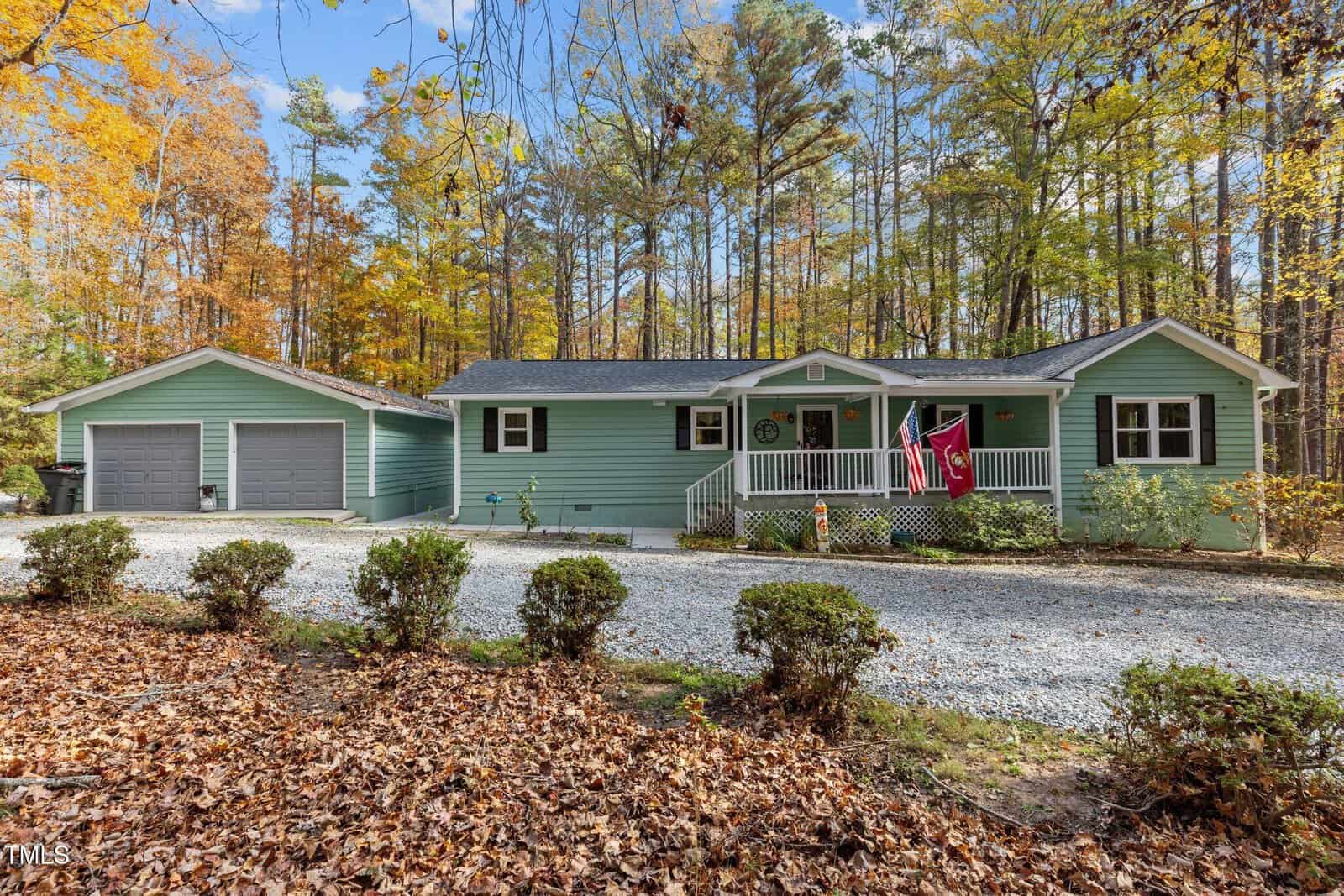 Located at 416 Forest Home Lane, this single-story green house boasts a welcoming front porch and a two-car garage. Surrounded by vibrant autumn trees in Henderson, it features flags near the entrance and a gravel driveway lined with shrubs and fallen leaves.