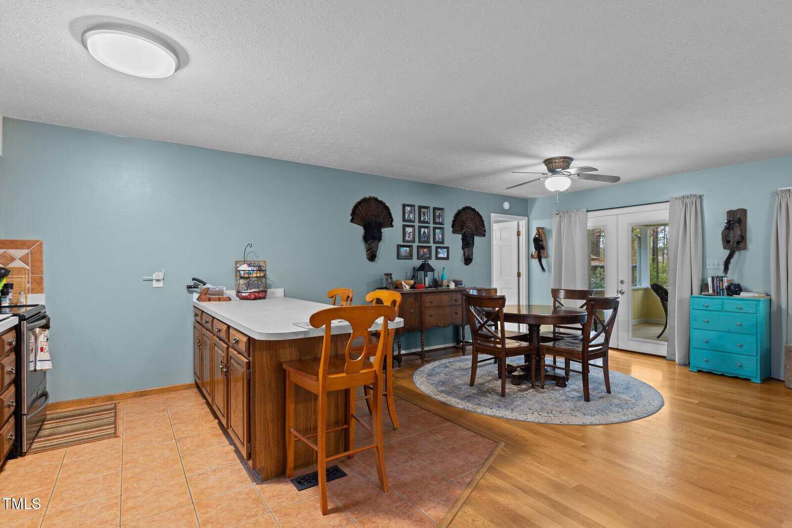 A cozy dining room with a round table and four wooden chairs sits on a circular rug. The walls are painted teal and decorated with framed pictures and mounted helmet displays. There are sliding glass doors leading to an outdoor area.