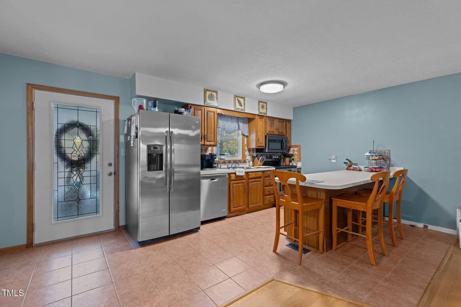 A kitchen with wooden cabinets, a stainless steel fridge, and a small island with three stools. There's a door with a decorative glass panel, light blue walls, and tiled floor. A window above the sink has a valance with a checkered pattern.