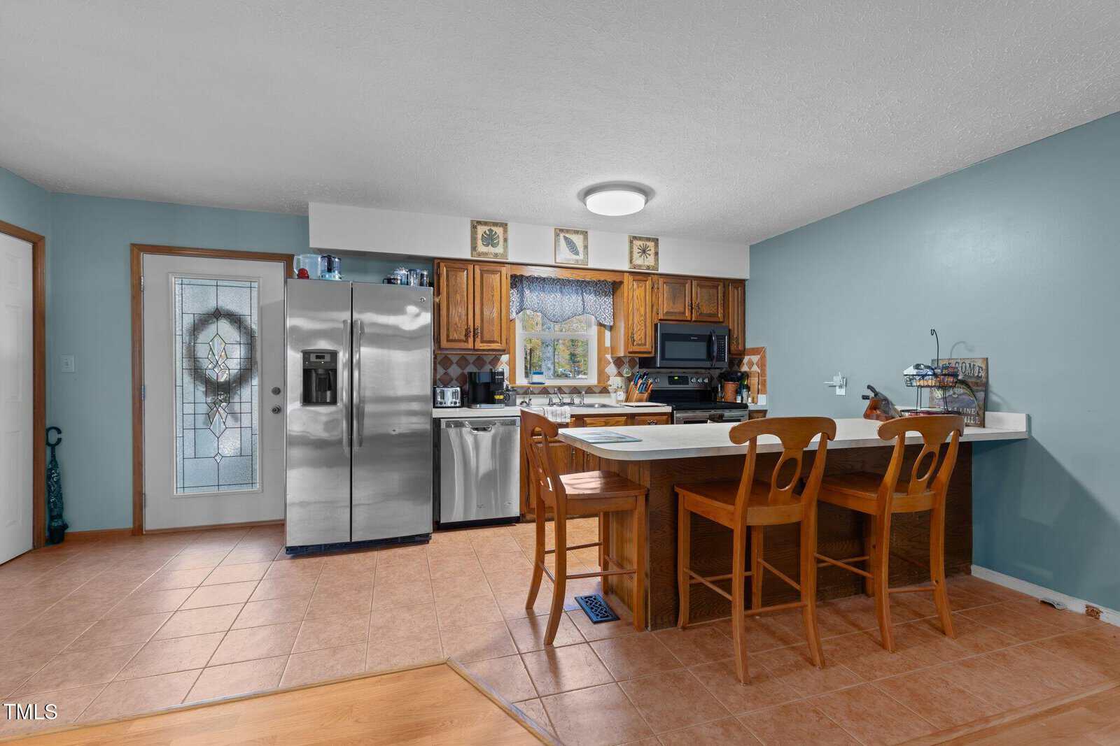 A kitchen with teal walls features wooden cabinets, a stainless steel fridge, and a dishwasher. A breakfast bar with wooden chairs faces the cooking area. A decorative glass door is on the left, and tile flooring spreads across the room.