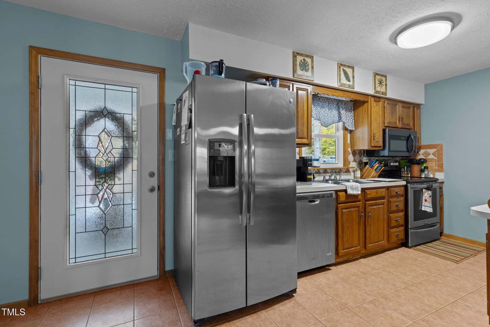 A kitchen featuring a stainless steel refrigerator, wooden cabinets, a dishwasher, and a microwave. A decorative glass door is on the left, and tiled floors and a backsplash complete the cozy look.