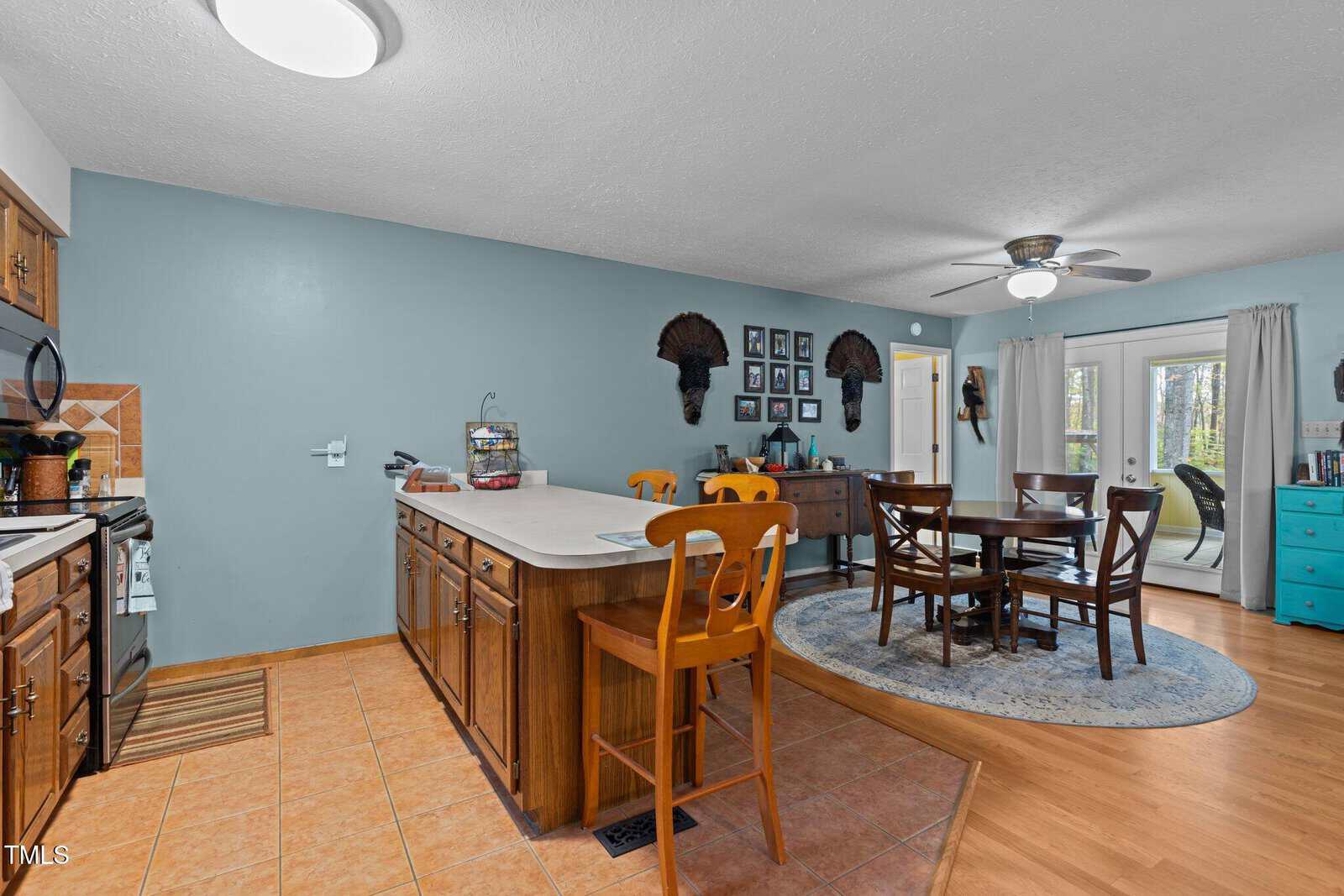 A cozy kitchen and dining area featuring an island with bar stools, wooden cabinets, and stainless steel appliances. A dining table with chairs sits on a round rug. Wall decor includes animal heads and framed pictures, with doors leading outside.