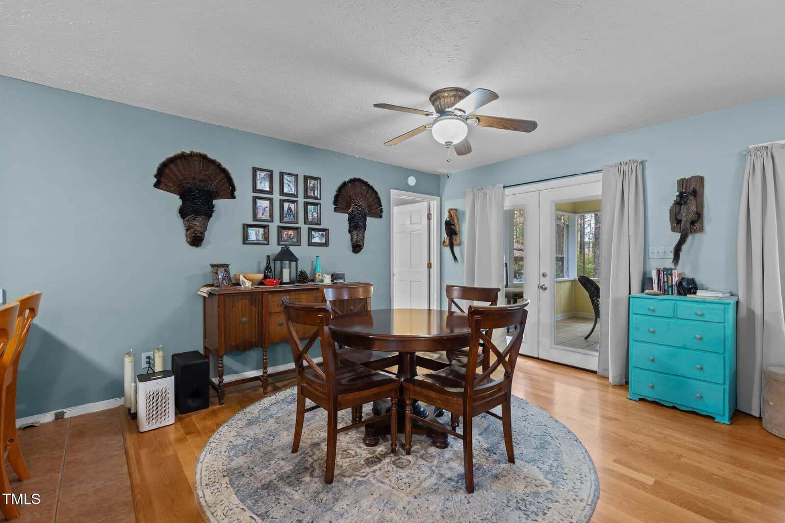 A dining room with a round wooden table and chairs on a circular rug. The walls are light blue with framed pictures and decorative items. Two turkey mounts are displayed on the wall. A white door with windows leads outside, and a teal dresser is on the right.