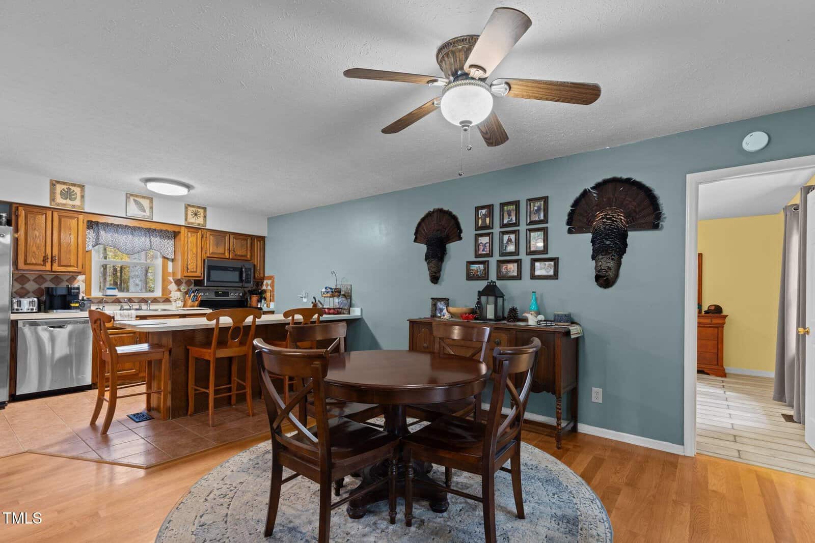 A cozy kitchen and dining area with wood flooring and a ceiling fan. The kitchen has wooden cabinets, a dishwasher, and a bar counter with stools. The dining area features a round table with chairs, a rug, and a wall decorated with framed pictures and decor.