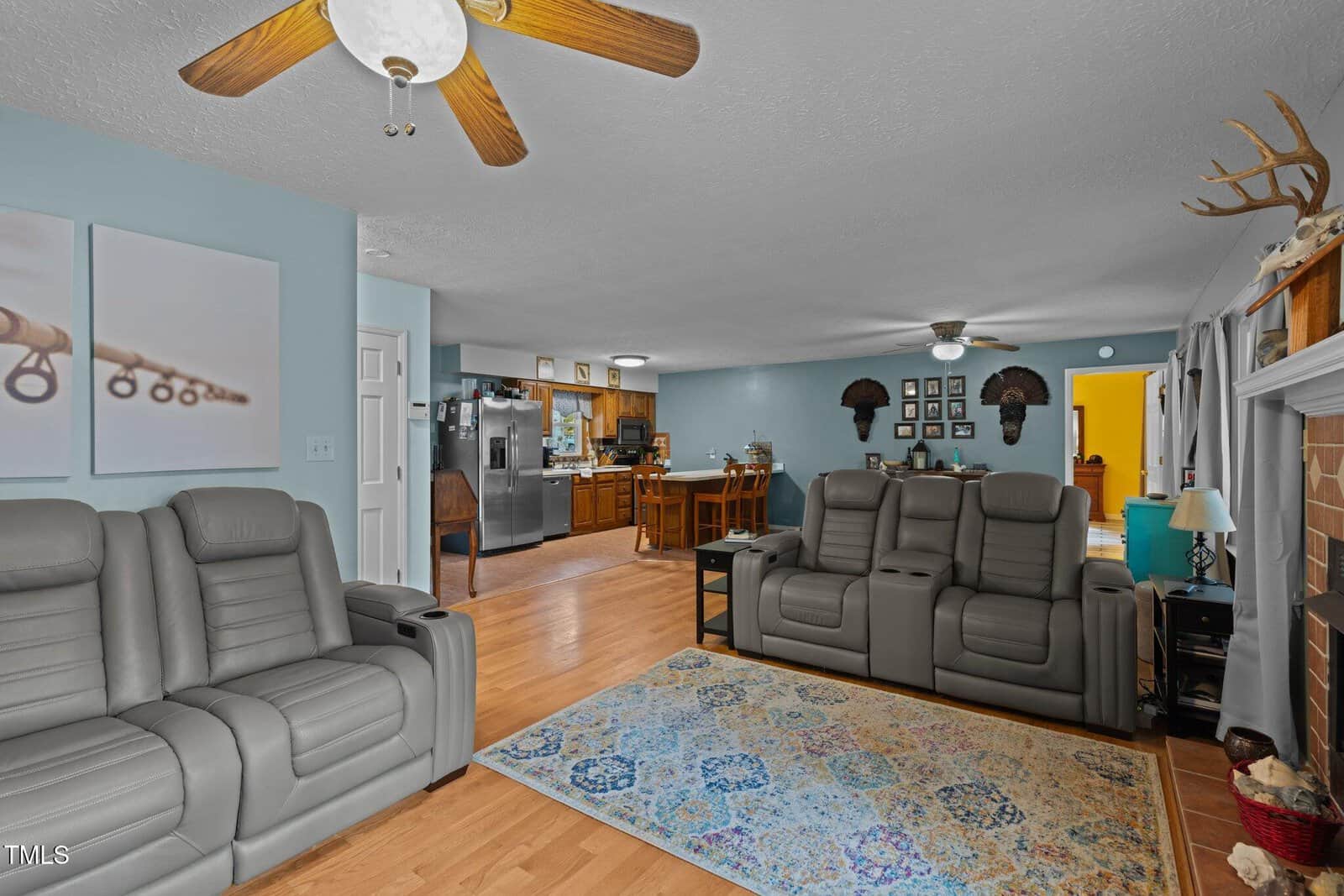 A spacious living room with two gray recliners on a light wood floor. A colorful rug adds warmth, and a ceiling fan hangs overhead. The room leads to an open kitchen and dining area. Walls are decorated with two paintings and mounted animal heads.