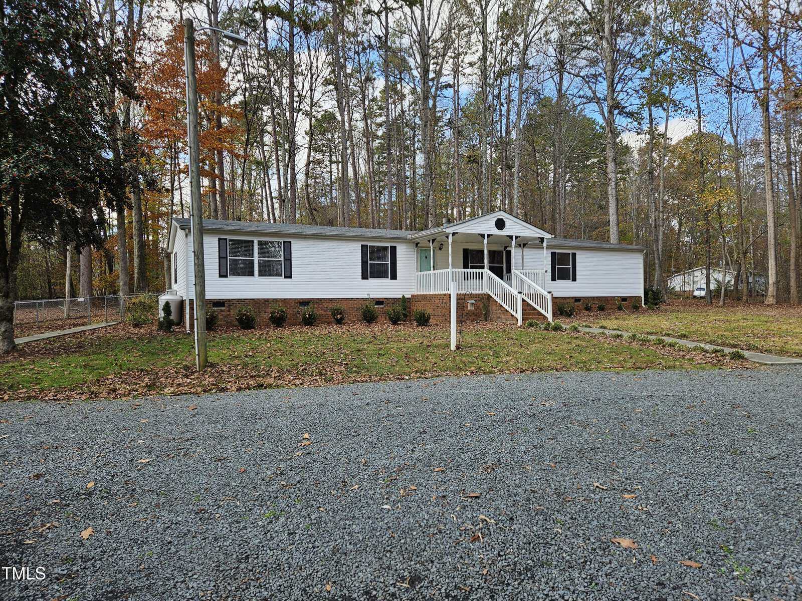 A white single-story house with gray shutters is set in a wooded area. A gravel driveway leads to the front door, which is accessed by a small porch with white railings. The surrounding trees are partially covered in autumn leaves.