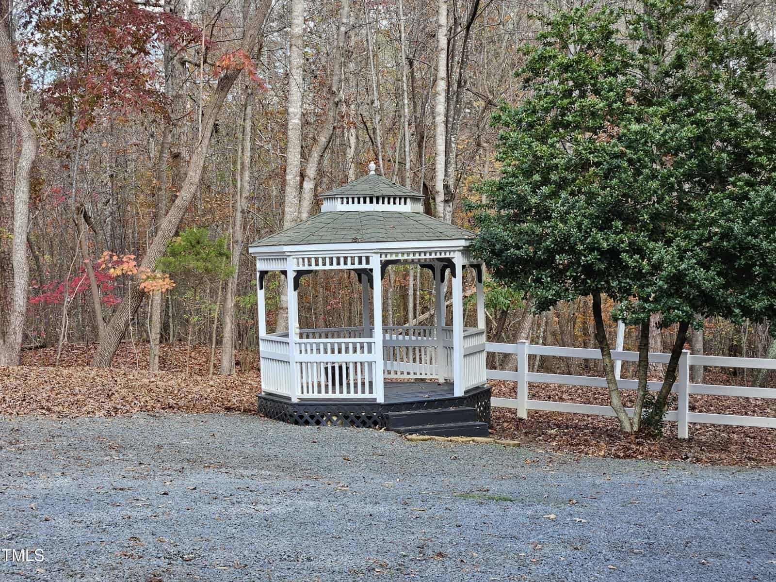 A white wooden gazebo stands in a wooded area with autumn leaves on the ground. A white fence is on the right and trees with bare branches are in the background. The ground is covered with gravel.